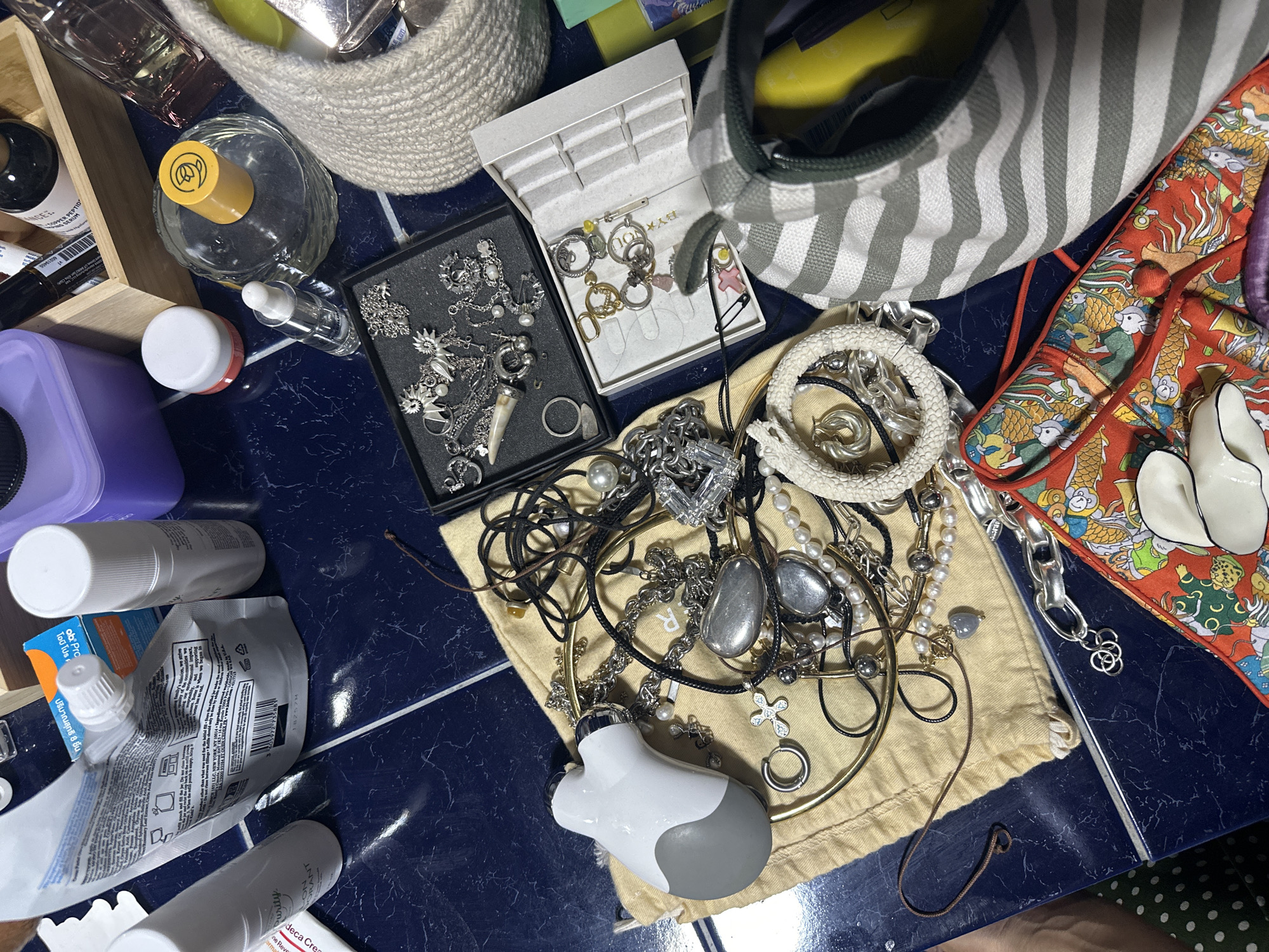 The image shows a collection of personal items, including jewelry, toiletries, and cosmetics, spread out on a dark blue tiled surface. In the foreground, a beige drawstring bag is filled with various necklaces, bracelets, and charms. To the left of the bag is a white and gray handheld electronic device. A black and white striped canvas bag is open in the lower right corner, revealing a yellow object inside. A patterned pouch with dragon motifs is also visible in the lower part of the image. In the background, there are several bottles and containers, including a clear glass perfume bottle with a yellow cap, a white spray bottle, and a purple container. The items appear to be arranged on a table in a domestic setting. There are no people visible in the image. The exact location in Baan Tai, Thailand, is not ascertainable from the provided image.
