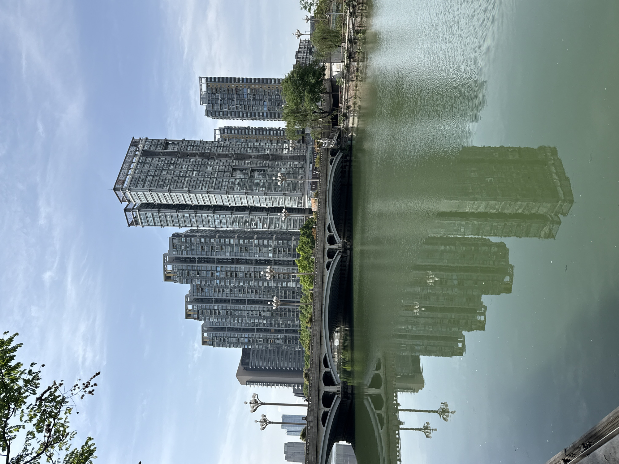 This daytime photograph, taken in Jinjiang District, China, captures a modern urban landscape alongside a body of water. The scene features a prominent row of high-rise buildings, a multi-arched bridge, and serene water with clear reflections, all under a bright sky.
On the left side of the frame, several tall, contemporary buildings stand vertically. These structures are characterized by their dark, reflective glass facades and numerous windows, suggesting they are residential or commercial towers. Above them, a clear, light blue sky stretches upwards, dotted with sparse, wispy white clouds, indicating sunny and pleasant weather conditions.
A distinctive multi-arched bridge spans across the water, providing an architectural focal point. The bridge, likely made of stone or concrete, features ornate streetlights lining its railing. Green foliage, including trees and bushes, can be seen along the riverbanks and near the bridge, adding natural elements to the urban setting.
The right side of the image is dominated by the calm, greenish-grey water, likely a river or canal. A striking feature of the photograph is the almost perfectly mirrored reflection of the buildings, the bridge, and the streetlights on the water's surface. These reflections, although slightly distorted by subtle ripples, create a strong sense of symmetry and depth.
No people or specific activities are visible in the scene, which presents a tranquil yet grand view of the cityscape. The time of day is clearly daylight, illuminated by natural sunlight. There is no discernible text visible within the photograph.