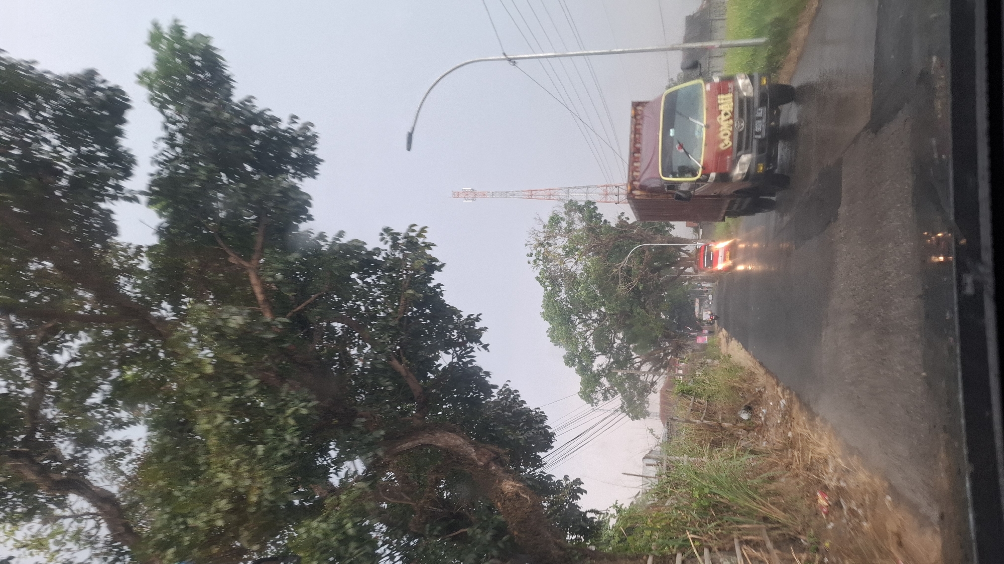 The image depicts a street scene in Sukasari, Indonesia, on a cloudy or overcast day, with evidence of recent rain given the wet road surface. On the left side of the road, there is overgrown vegetation and a wooden fence. Further down the road, there are buildings. A street lamp is visible on the right side of the road, with overhead power lines. A tall cell tower is also visible in the distance, painted with orange and white sections.
In the foreground, a red truck with a yellow stripe and the word "GOMBALL" written on its side is driving on the right side of the road. The truck has a dark-colored cargo box covered with a tarp. Its headlights are on. Behind the truck, a red car is also visible, its taillights illuminated and reflecting on the wet road. There appear to be other vehicles further down the road, but they are indistinct. The image is taken from inside a vehicle, likely looking through the windshield, as evidenced by the reflection of lights at the bottom of the frame. A large tree with thick branches and green leaves dominates the upper left portion of the image, casting shadows.