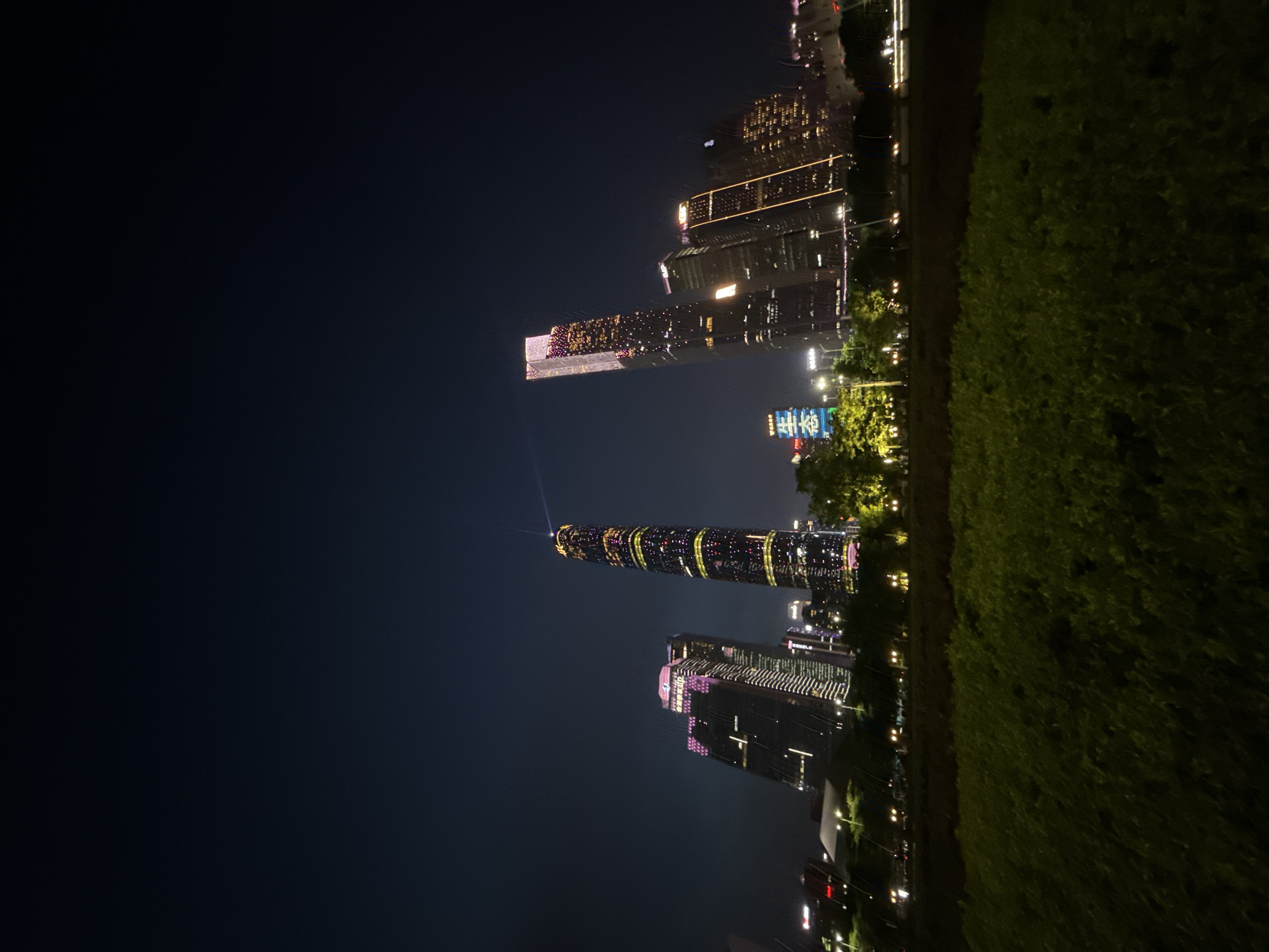The image shows a nighttime view of the Guangzhou International Finance Center and other skyscrapers in the Tianhe District of Guangzhou, China. The buildings are illuminated with colorful lights, creating a vibrant urban landscape. In the foreground, a grassy area is visible, with streetlights and trees lining the path. The sky is dark, indicating it is nighttime.