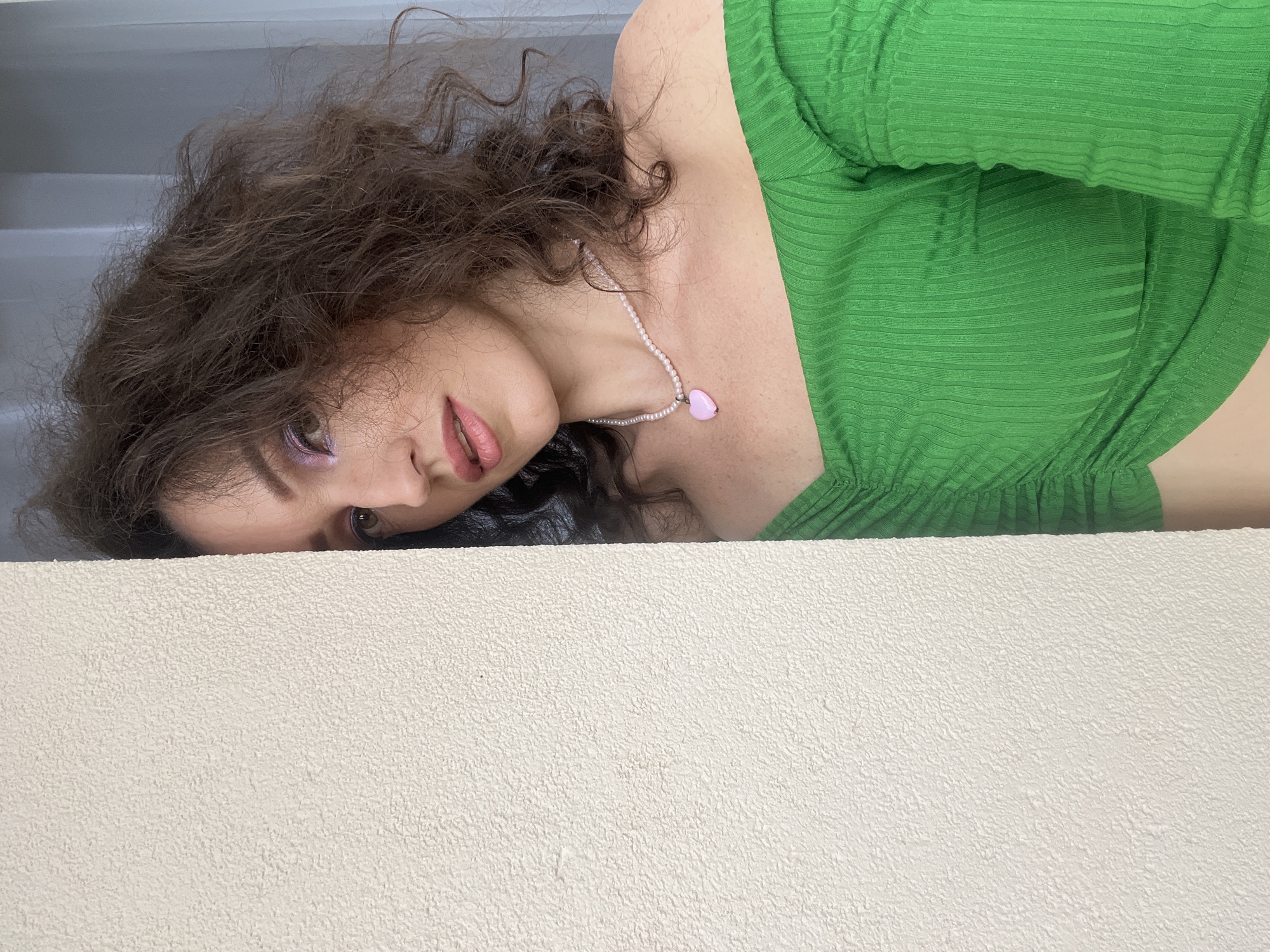 A woman with curly brown hair and pink lipstick leans against a textured, cream-colored wall. She is wearing a bright green, off-the-shoulder top and a pearl necklace with a pink heart pendant. Her eyes are made up with purple eyeshadow. The background includes a light blue sheer curtain. The scene is indoors.
