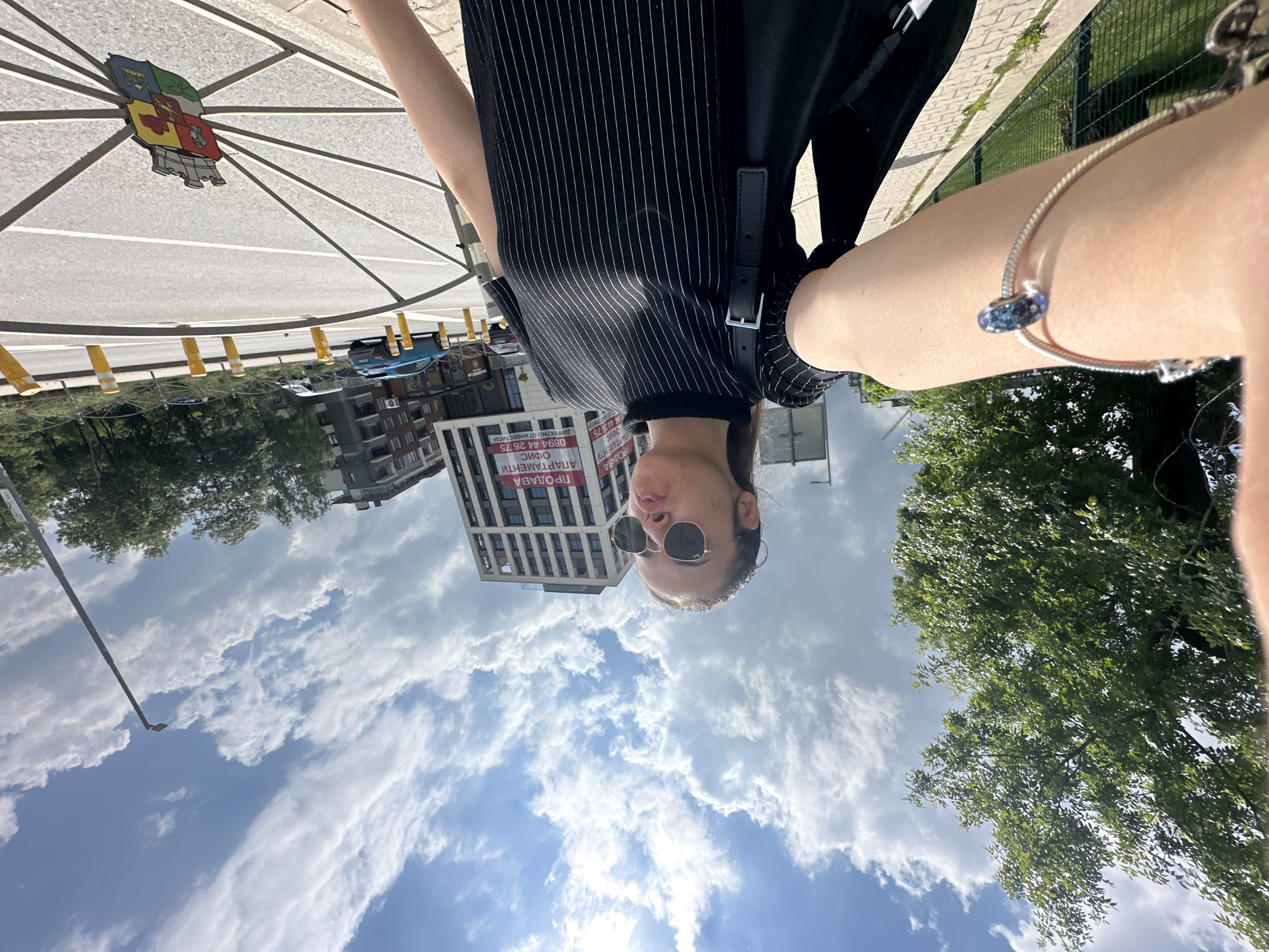 This unique image presents an inverted perspective, appearing as if the ground is above and the sky below, likely a result of an upward-facing selfie or a deliberate photographic choice.
In the foreground, a person is visible, wearing dark sunglasses and a black top with thin white vertical stripes. Their arm, adorned with a silver snake-chain bracelet featuring a blue, iridescent bead charm, is extended towards the camera. The individual appears to be taking a selfie, with their head tilted back and looking up (or down, given the inversion).
The scene captures an urban environment under bright daylight. What would typically be the ground is at the top of the frame, showing a light-colored, possibly concrete or tiled surface with a distinctive large circular design featuring spokes radiating from a central, colorful emblem resembling a coat of arms or crest. Along the side of this "ground," yellow bollards line what appears to be a street, where parts of vehicles are faintly visible.
In the background, several buildings are present, including a taller, modern-looking structure. A large banner on one of the buildings displays Cyrillic text: "ПРОДАВА АПАРТАМЕНТИ ОФИС 0894 44 26 75 ДИРЕКТНО ОТ ИНВЕСТИТОР," which translates to "FOR SALE APARTMENTS OFFICE 0894 44 26 75 DIRECTLY FROM INVESTOR." This text, along with the provided context, indicates the location is Sofia, Bulgaria.
Lush green trees frame parts of the scene, and a green mesh fence is visible on the right. In the "sky" area at the bottom of the frame, scattered white cumulus clouds are prominent against a clear blue sky, with a single street lamp post extending into the view. The overall impression is a casual, creatively composed photograph taken during the daytime.