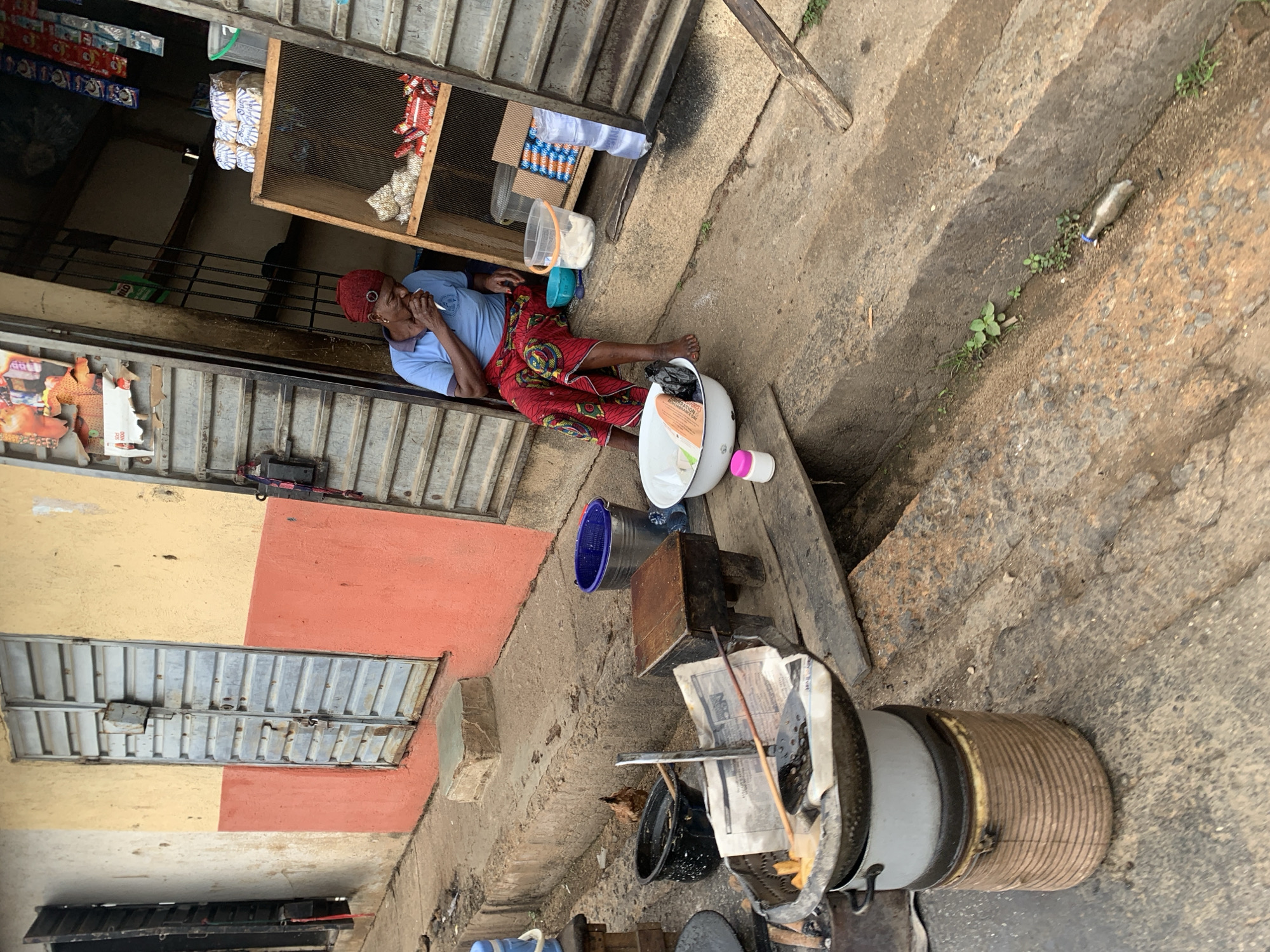 An elderly woman with a red head covering lies on a concrete walkway in front of a shop in Jos, Nigeria. The shop is partially open, displaying assorted goods. The woman is wearing a blue shirt and a red patterned skirt. Her right hand is near her mouth. Next to her is a white basin, a blue bucket, and a wooden box. In the foreground, there is a metal cooking pot with a coiled exterior, a black bucket, and other cooking utensils. The ground is concrete and appears worn. The buildings are painted yellow and orange.