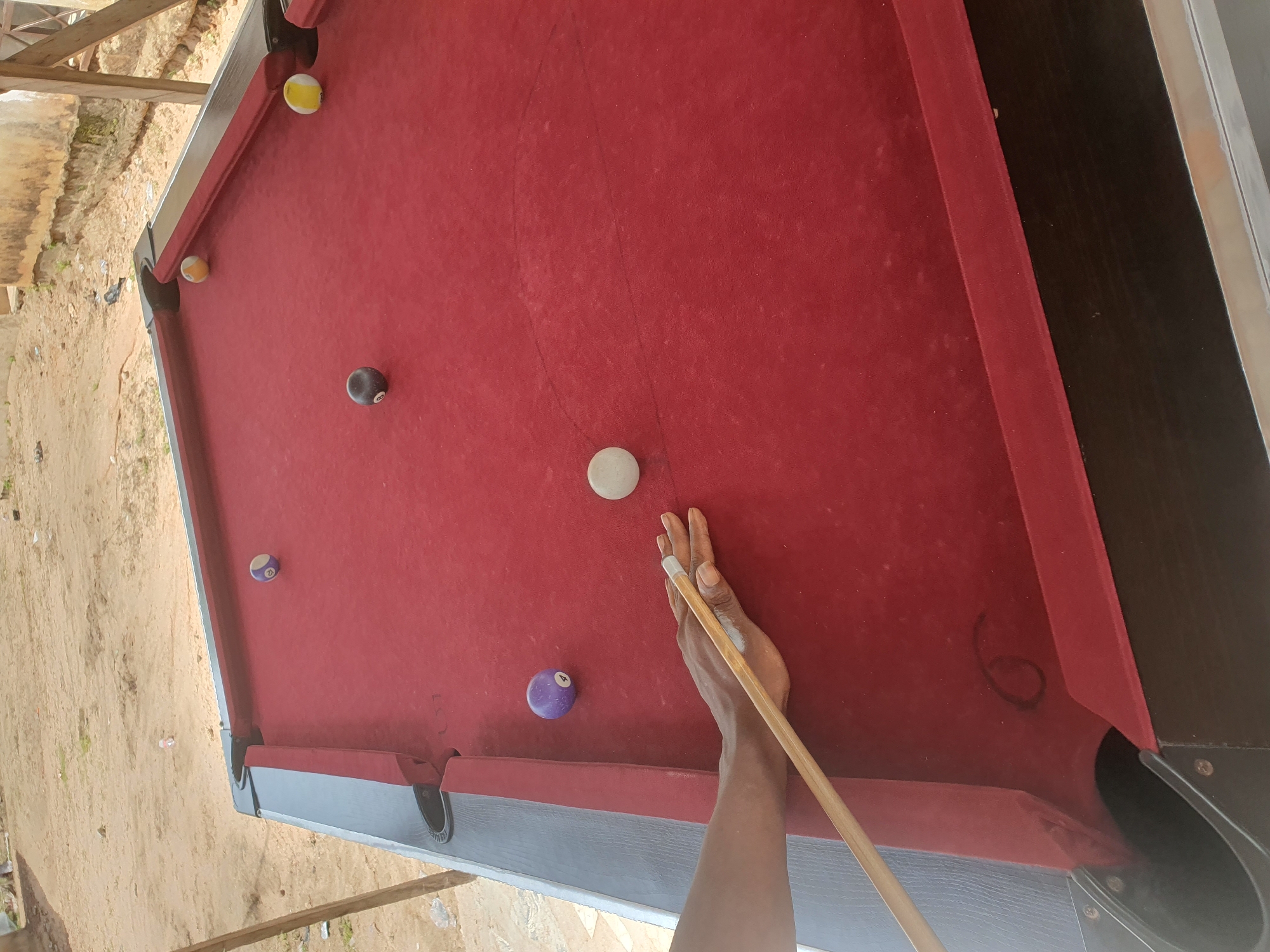 In Lokoja, Nigeria, a person's hand is visible, holding a pool cue and poised to strike a white cue ball on a red felt billiard table. The table has several billiard balls scattered across its surface: a purple ball with the number 4, a dark-colored ball with the number 8, and two striped balls, one yellow and white, and another with a yellow and white pattern with a blue stripe. The background reveals a sandy outdoor environment with a weathered building and some vegetation, suggesting the billiard table is situated in an open-air or partially covered area.