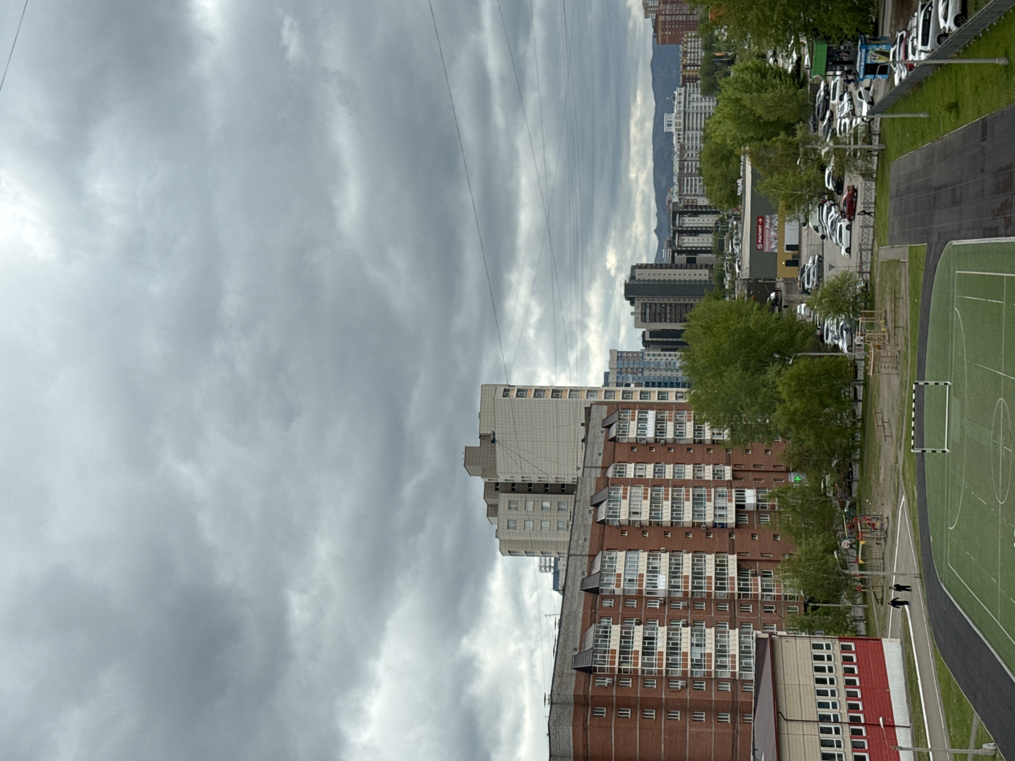 The image depicts a cityscape of Krasnoyarsk, Russia, under a cloudy sky. The foreground features a multistory apartment building with brick and white facade sections. Adjacent to the building is a school playground and running track. In the middle ground are additional apartment buildings, and in the distance, there is a view of mountains behind the city's buildings. Power lines are visible overhead. On the right of the image there is a line of parked cars on a street lined with trees. Two figures appear to be walking along the path.