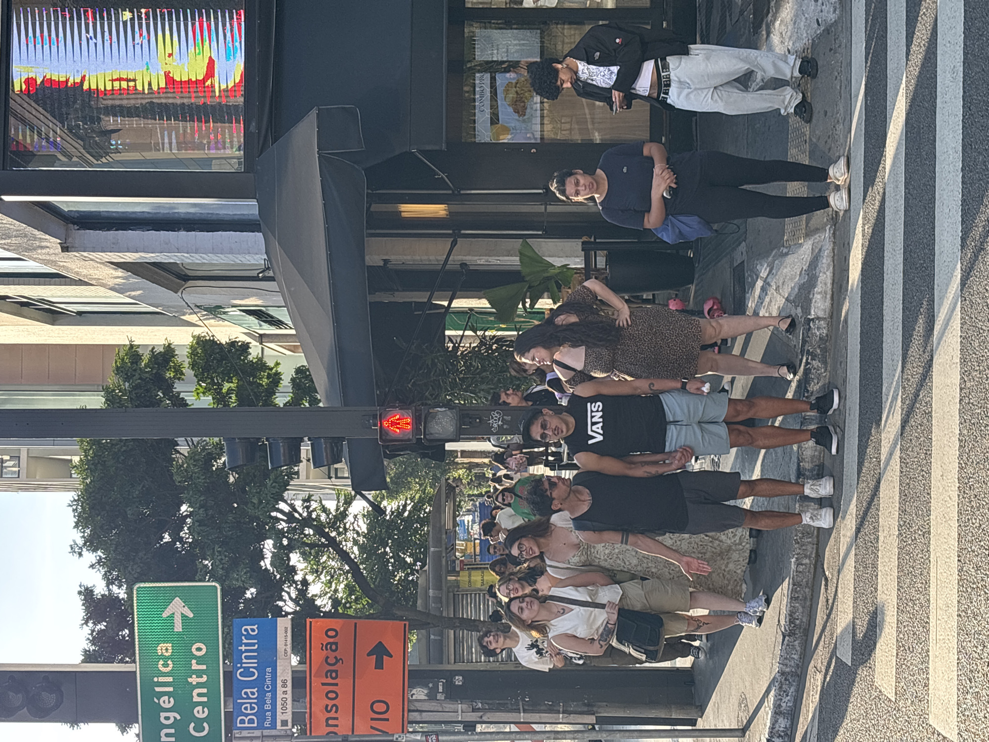 The image captures a group of people standing on a zebra crossing at a street intersection in São Paulo, Brazil. The location is suggested by street signs indicating "Angélica Centro" and "Consolação VIO," pointing in opposite directions. Another sign references "Bela Cintra, Rua Bela Cintra, 1050 a 86."
A red traffic light is visible. The group of people appears to be waiting to cross the street. Among them, one person is wearing a "Vans" shirt. Some individuals are engaged in conversation with each other. Several businesses with awnings are present along the street. The sidewalks appear busy.
