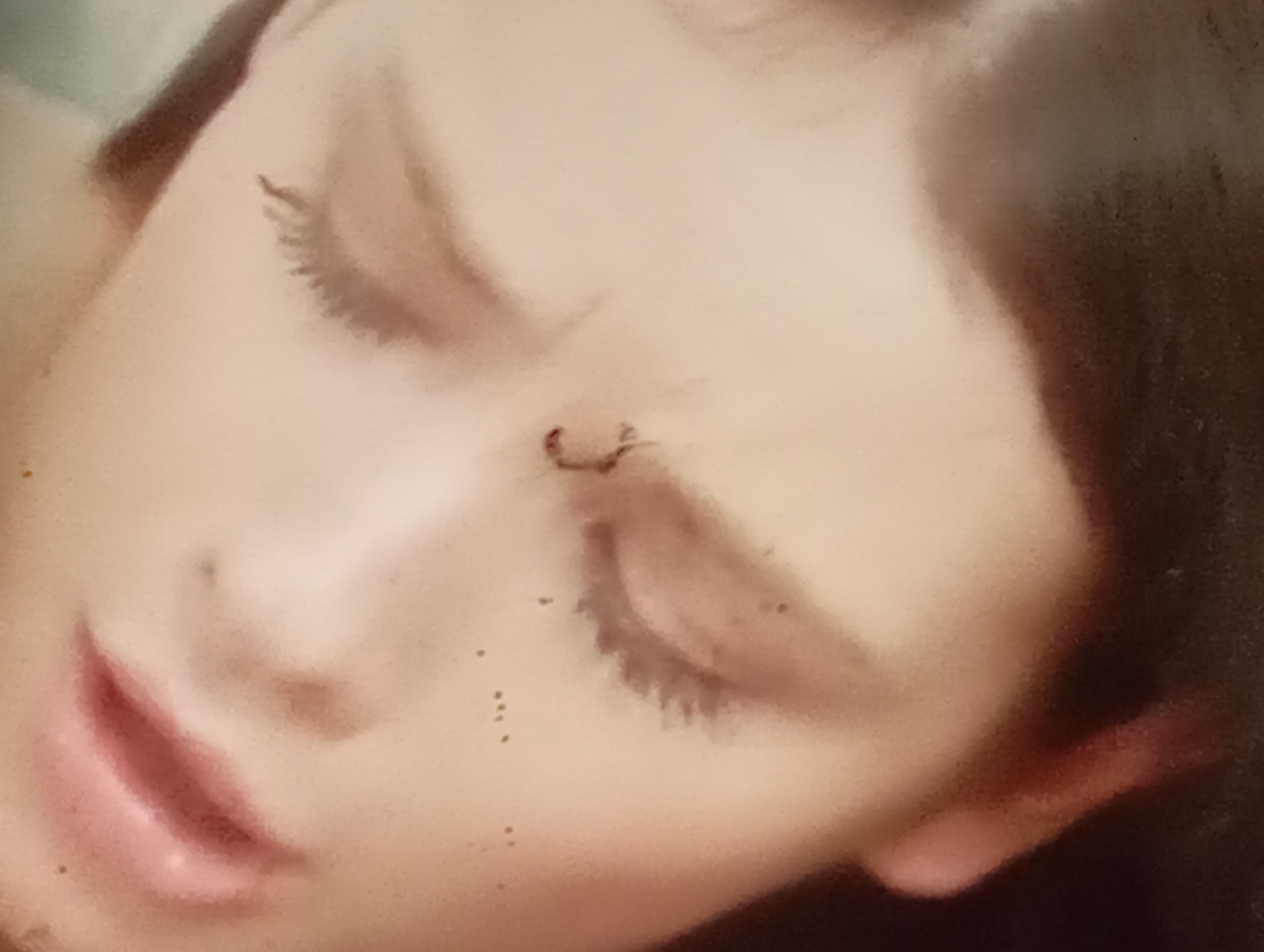 The media file shows a close-up, blurry image of a person's face, specifically focusing on their eye and lip area. The person appears to be female, with dark hair visible on the right side of the frame. Their eyes are closed, and their eyelashes are long and dark. There is a small, dark marking or piece of debris on the bridge of their nose, just below the eye. The lips are slightly parted, revealing a soft pink color. The overall impression is one of rest or sleep. The lighting is soft and diffused, suggesting an indoor setting or possibly a cloudy day. There are several small dark specks scattered across the skin, which could be dust or blemishes. The background is out of focus and appears to be a light, neutral color. No text is visible in the image. The subject's facial expression, with closed eyes, suggests a state of relaxation or unconsciousness.