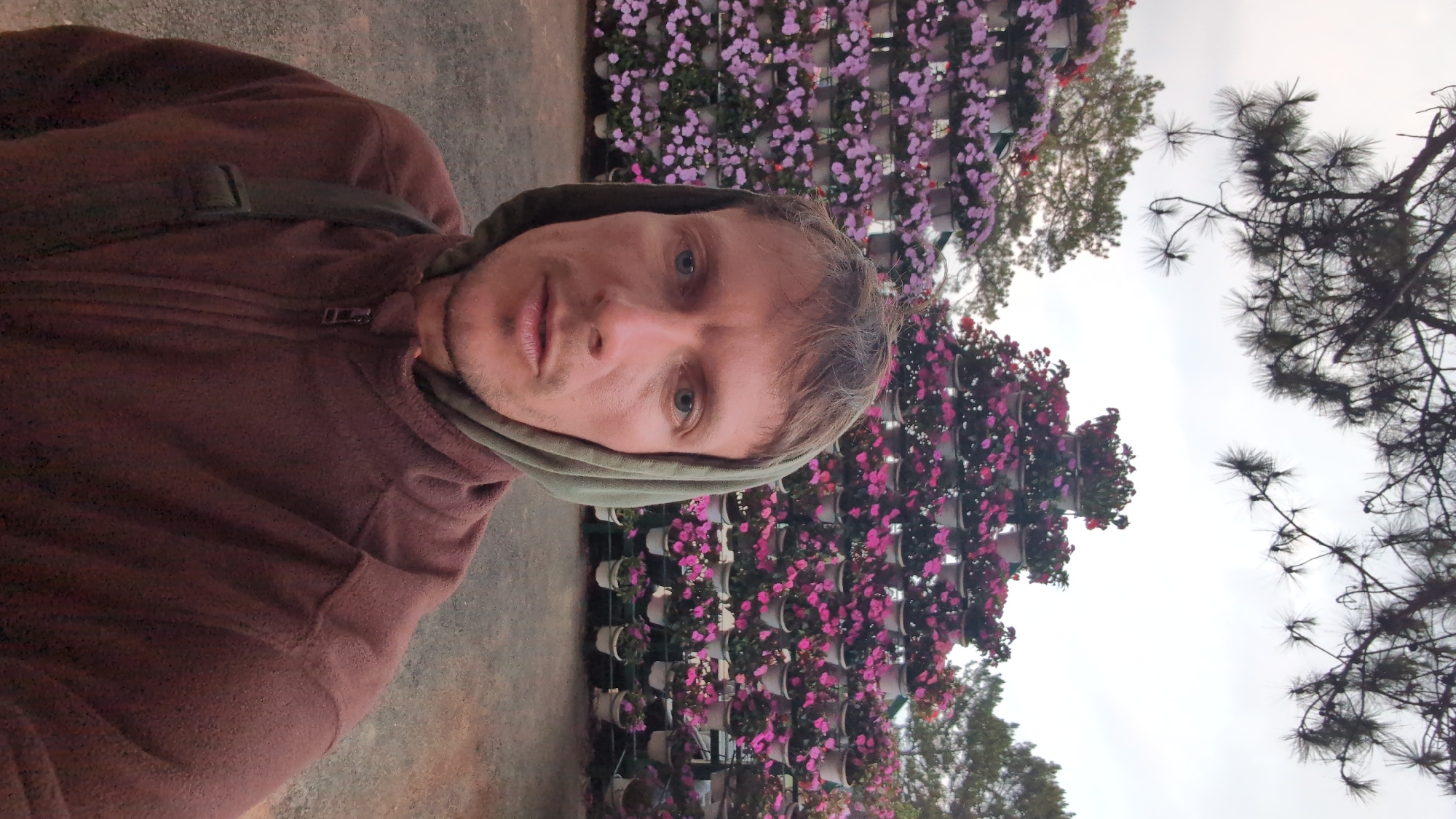 A man is taking a selfie in front of a flower wall in Da Lat, Vietnam. The man is wearing a brown fleece jacket with the hood up. Behind him is a wall of flowers in pots. There are also trees visible in the background. The sky is overcast.
