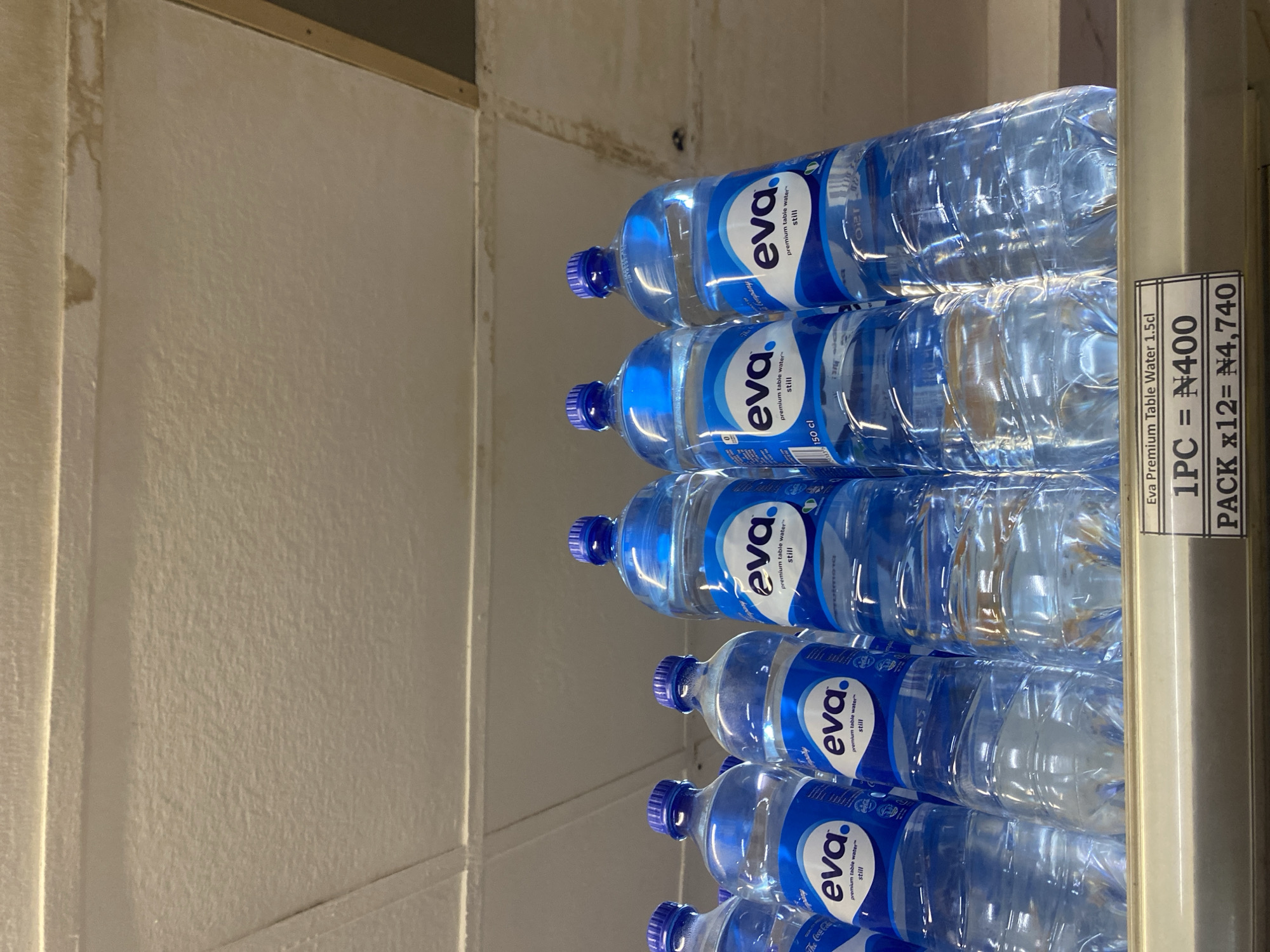 The image shows a shelf with multiple bottles of Eva premium table water. The bottles are clear plastic with blue caps and feature blue and white labels. A price tag is visible at the bottom of the shelf, indicating "Eva Premium Table Water 1.5cl", "1PC = N400", and "PACK x12 = N4,740". The background shows a textured ceiling. This scene is likely from a retail store in Ikorodu, Nigeria, given the currency symbol (N for Naira) and the context of bottled water being sold.