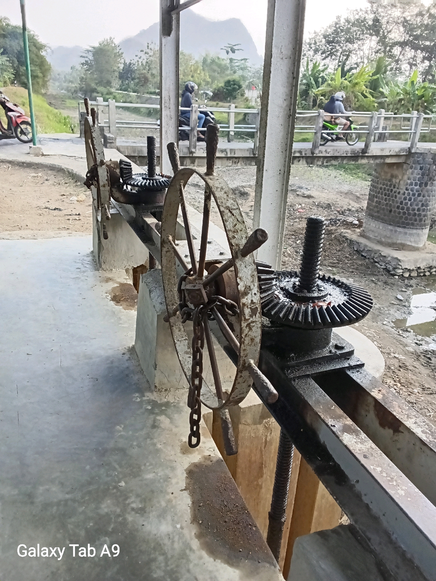 The image depicts a water control gate mechanism at the irrigation canal in Sumberingin, Indonesia. In the foreground, a large, rusty metal wheel, part of the gate's operating system, is prominently featured. It is connected to a series of gears and a threaded rod, all showing signs of wear and lubrication. The mechanism is mounted on concrete supports.
In the background, a concrete bridge spans the canal, with a white railing. Two people on motorcycles are visible on the bridge. One motorcycle is red, and the other is green. Lush green foliage and trees are abundant on either side of the canal and bridge, with a hazy mountainous landscape in the distance. The ground around the canal appears to be a mix of dirt and concrete. The sky is overcast, suggesting possibly diffused light conditions. A watermark in the bottom left corner reads "Galaxy Tab A9".