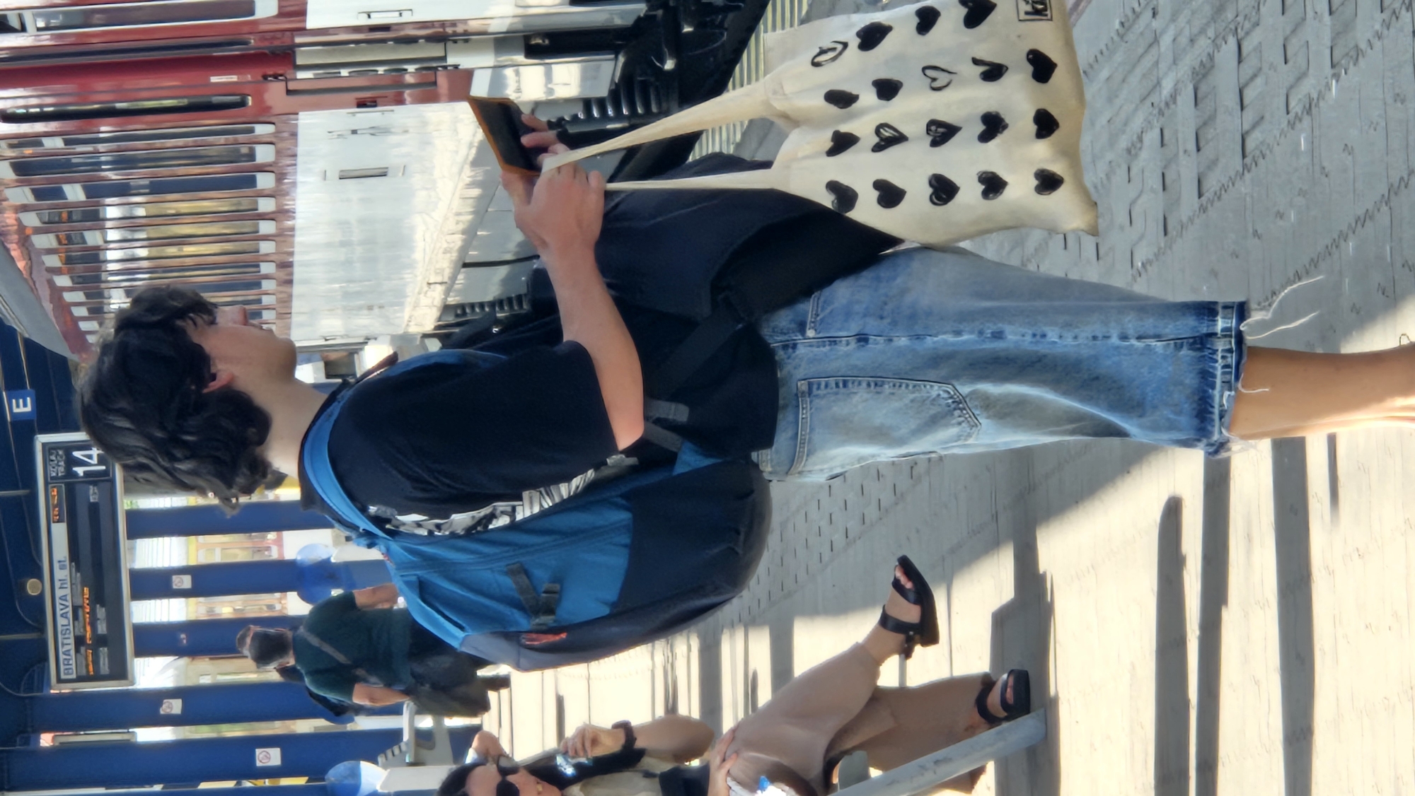 The image captures a scene at the Bratislava hlavná stanica (main train station) in Bratislava, Slovakia. A young person with dark, wavy hair is standing on the platform, holding a mobile device and a tote bag decorated with black hearts. They are wearing a black t-shirt and denim shorts, with a blue backpack slung over one shoulder. To the left, a woman with dark hair is seated on a bench, holding an object in her hand. Another person, wearing a green t-shirt and a dark bag, is walking in the background. The train station platform is visible, with track numbers and station information displayed on a sign. The sun casts long shadows on the wooden platform.
