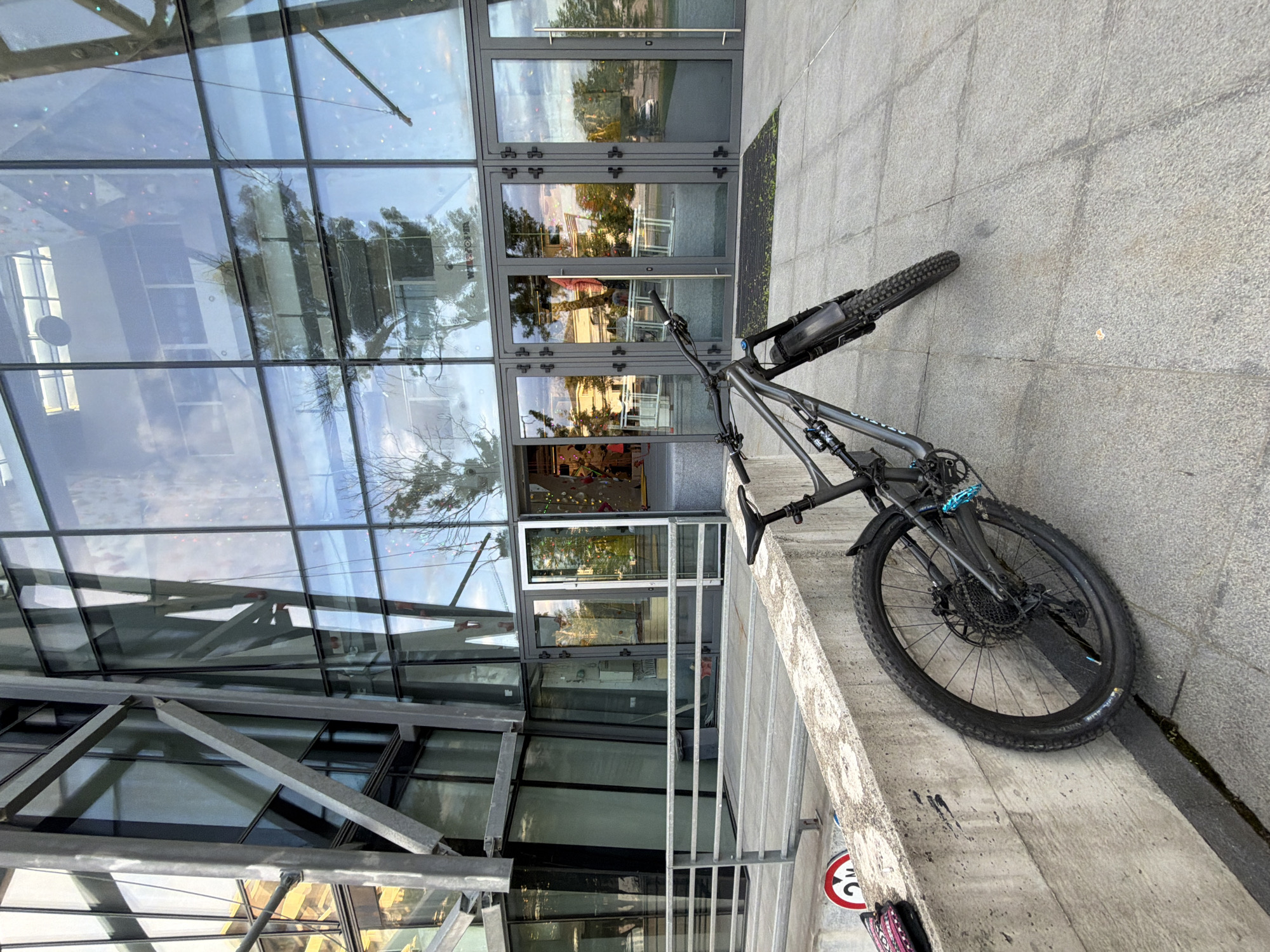 A full-suspension mountain bike, dark grey with subtle blue accents, is parked leaning against a concrete barrier next to a modern building with a striking glass facade. The image is taken from a slightly elevated perspective, looking down and across the scene.
The bike features knobby tires, disc brakes, and a frame bag attached to the top tube. Part of the brand name "SPECIALIZED" is visible on the downtube. Its front wheel rests against the concrete wall, while the rear wheel is on the tiled pavement.
The building behind the bike is composed almost entirely of large, rectangular glass panels framed by dark metal. These panels reflect the surrounding environment, including a clear sky, bare tree branches, and distant trees with green foliage. Some reflections suggest indoor elements like colorful, small lights or decorations. Several double glass doors, also with dark metal frames, serve as the entrance. Below the glass facade, there are internal metal support beams visible through the windows. On one of the glass panels, the text "EUROGLASS" is partially legible.
The ground consists of grey tiled pavement. In the lower right corner, a red and white circular sign, typical for "no parking" or time-limited parking, is partially visible. The number "30" can be seen on this sign. The time of day appears to be daytime, possibly late afternoon, given the quality of light and reflections. The setting is an urban environment in Sofia, Bulgaria.
