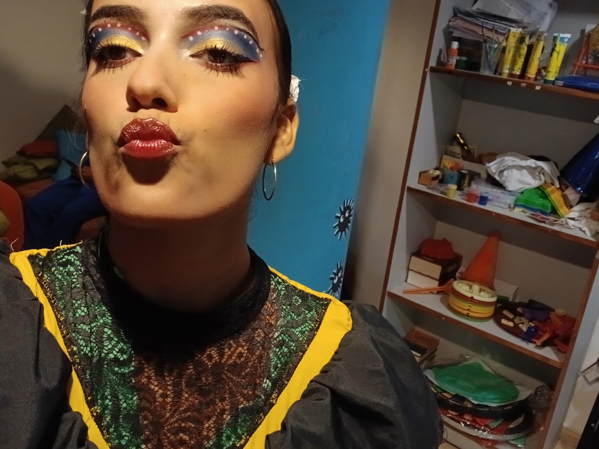 A person is posing for a selfie indoors. The individual has fair skin, brown eyes, and dark hair pulled back. They are wearing makeup, including yellow and blue eyeshadow with small white dots, and red lipstick. They are pouting their lips for the camera. The person is wearing hoop earrings and a black and yellow top with lace detailing. Behind the person is a blue wall with dark blue snowflake-like decals. To the right of the person is a white shelving unit with various objects on the shelves, including books, containers of art supplies, and a cone. There is no explicit indication the location is Playa El Agua, Venezuela in the image.