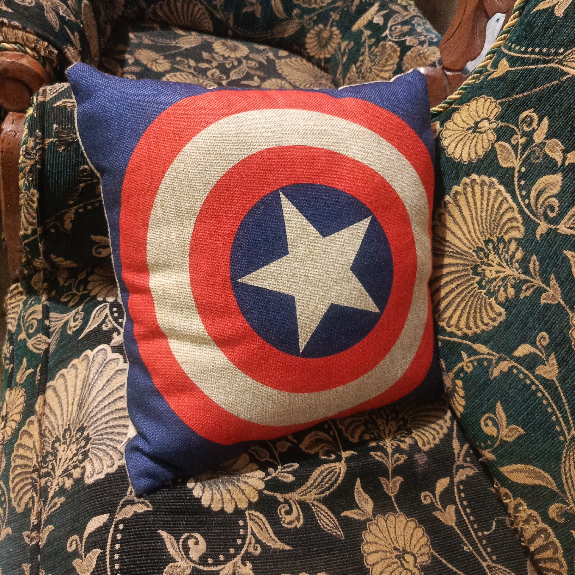 A close-up, eye-level shot captures a Captain America shield-patterned throw pillow placed on a dark green chair with a gold floral pattern. The pillow is square and has a textured fabric. Its design consists of concentric circles in blue, red, and white, with a white star in the center of a blue circle. The chair's upholstery features a repeating motif of stylized flowers and leaves. The lighting appears to be indoor ambient light, casting soft shadows and highlighting the textures of the fabrics. There are no people or actions depicted in the image, and no specific location information such as Karangtuang, Indonesia is visible or inferable from the scene.