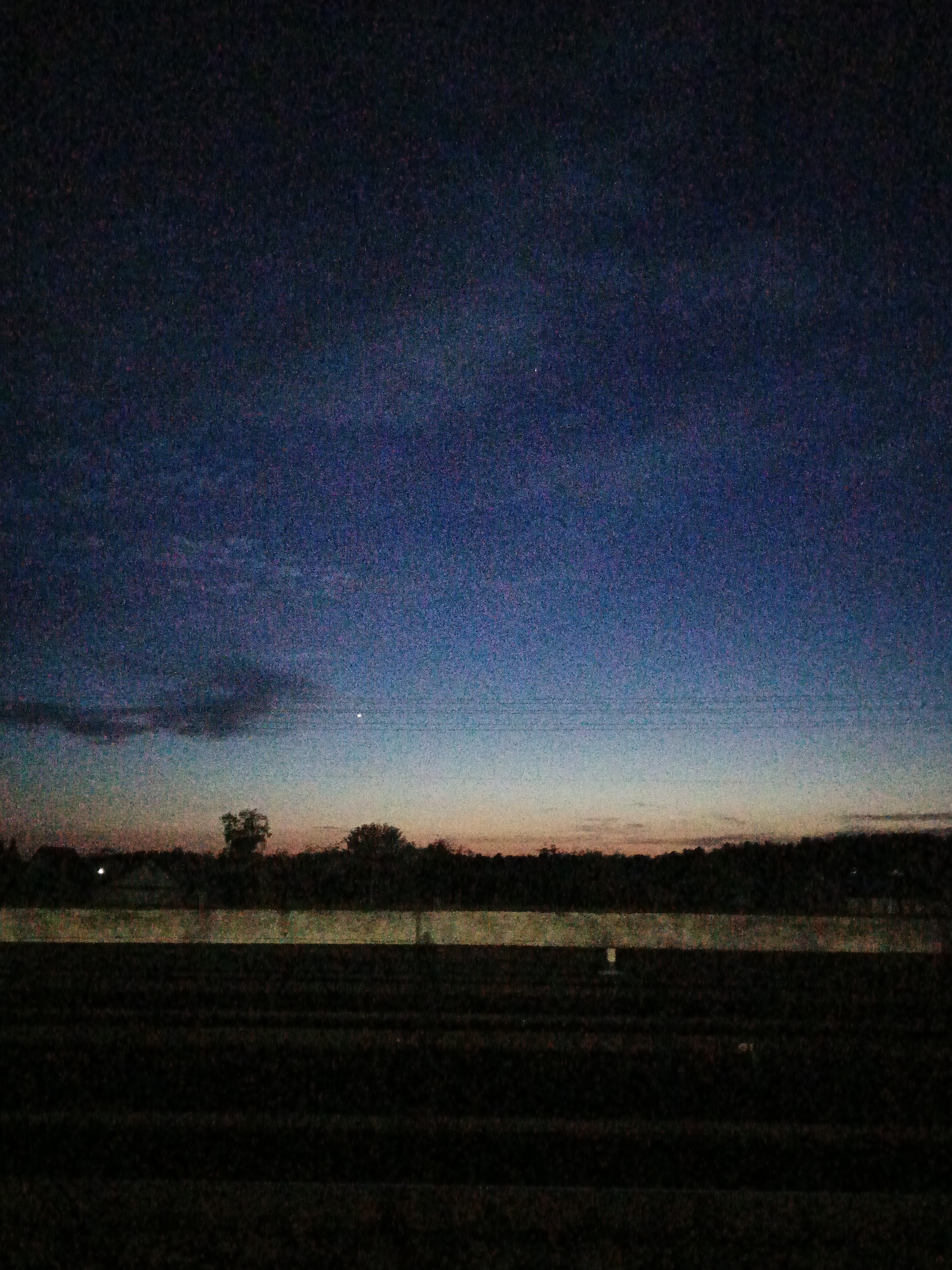 The image depicts an agricultural field in Лосева, Belarus at dusk or dawn. The sky transitions from a deep blue at the top to lighter shades of orange and pink near the horizon. A single bright point of light, possibly a star or planet, is visible in the sky. Silhouetted trees and a low-lying hedge or wall are present on the horizon, separating the field from the sky. The field itself is dark, with discernible rows of crops. The overall image is grainy and has low lighting conditions.