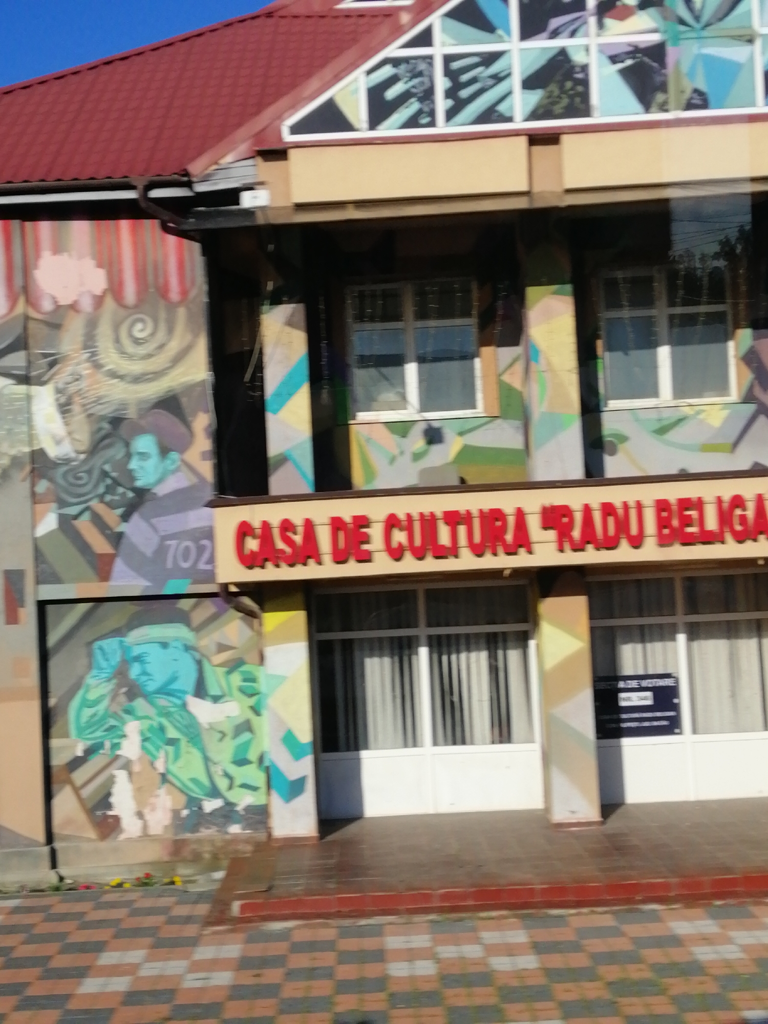 The image shows the exterior of a building adorned with vibrant murals. The building has a red tiled roof and several windows. A prominent sign with red, three-dimensional lettering reads "CASA DE CULTURA RADU BELIGA". The murals depict stylized portraits of people in what appears to be a somewhat abstract and geometric style, with strong color contrasts. One mural shows a figure in a hat with the number "702" visible on their clothing. Another mural features a figure wearing a military-style uniform. The building's facade is a mix of painted sections and windows, with colorful geometric patterns integrated into the design. In front of the building, a checkered tiled walkway is visible. The lighting suggests it is daytime, and the sky is clear blue. There is a black sign mounted on the building's exterior near a window, but the text on it is too blurry to read. The overall impression is that of a cultural center with an artistic and modern facade.