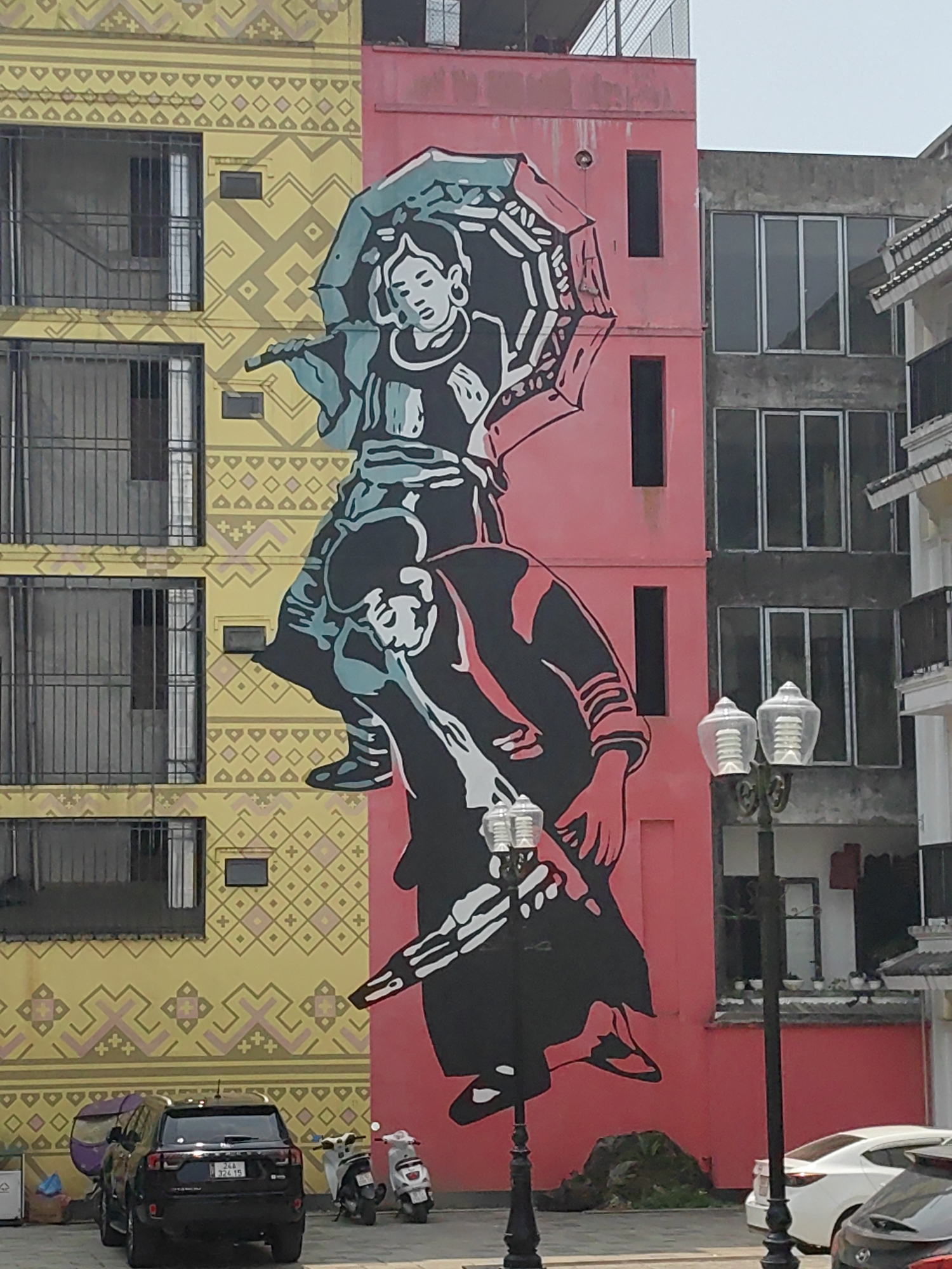The image displays an urban street scene in Sa Pa, Vietnam, centered on a large mural covering the side of a multi-story building. The mural features two stylized figures in traditional attire, rendered in black, teal, white, and red. The upper figure, a female, holds an open umbrella overhead, while the lower figure, a male, is depicted in a dynamic pose below her. This mural primarily covers a pink-painted section of the building and extends onto an adjacent yellow section decorated with repetitive geometric patterns.
The yellow building section to the left features multiple stories with window openings and dark metal balcony railings. The pink section serving as the mural's background contains several narrow, vertical rectangular windows. To the right, an additional multi-story building is visible, characterized by a grey concrete facade with large windows and a white section with a tiled roof overhang.
At ground level, a paved area serves as a street or parking lot. A black SUV with Vietnamese license plate "24A 326.15" is parked facing away from the viewer. Two scooters or motorbikes are parked behind the SUV. Further right, a white sedan and a dark-colored vehicle are partially visible. Two ornamental black street lamps with clear, ribbed plastic light covers stand prominently in the foreground on the right. A small patch of dark green foliage or rocks is situated at the base of the pink building. The scene is illuminated by daylight under a clear sky.