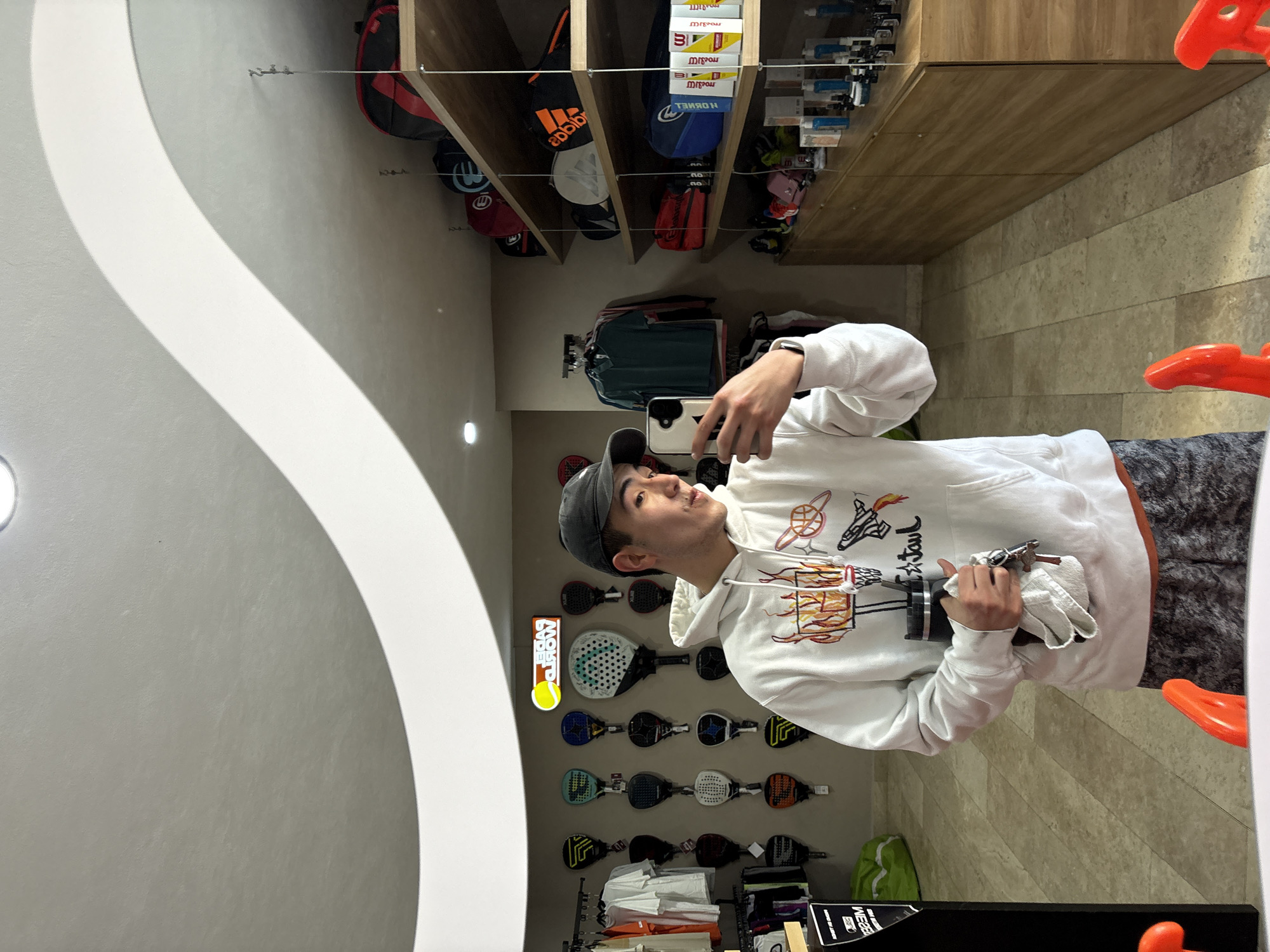 A mirror reflection shows a person wearing a white hoodie, a gray cap, and patterned gray pants holding a phone to take a selfie. The reflection reveals a sporting goods store with padel rackets displayed on the wall. Bags and boxes are stored on wooden shelves above. Clothes are hanging on a rack in the store. Orange chair elements are also visible in the mirror's reflection. The scene is located in San Bernardino Tlaxcalancingo, Mexico.