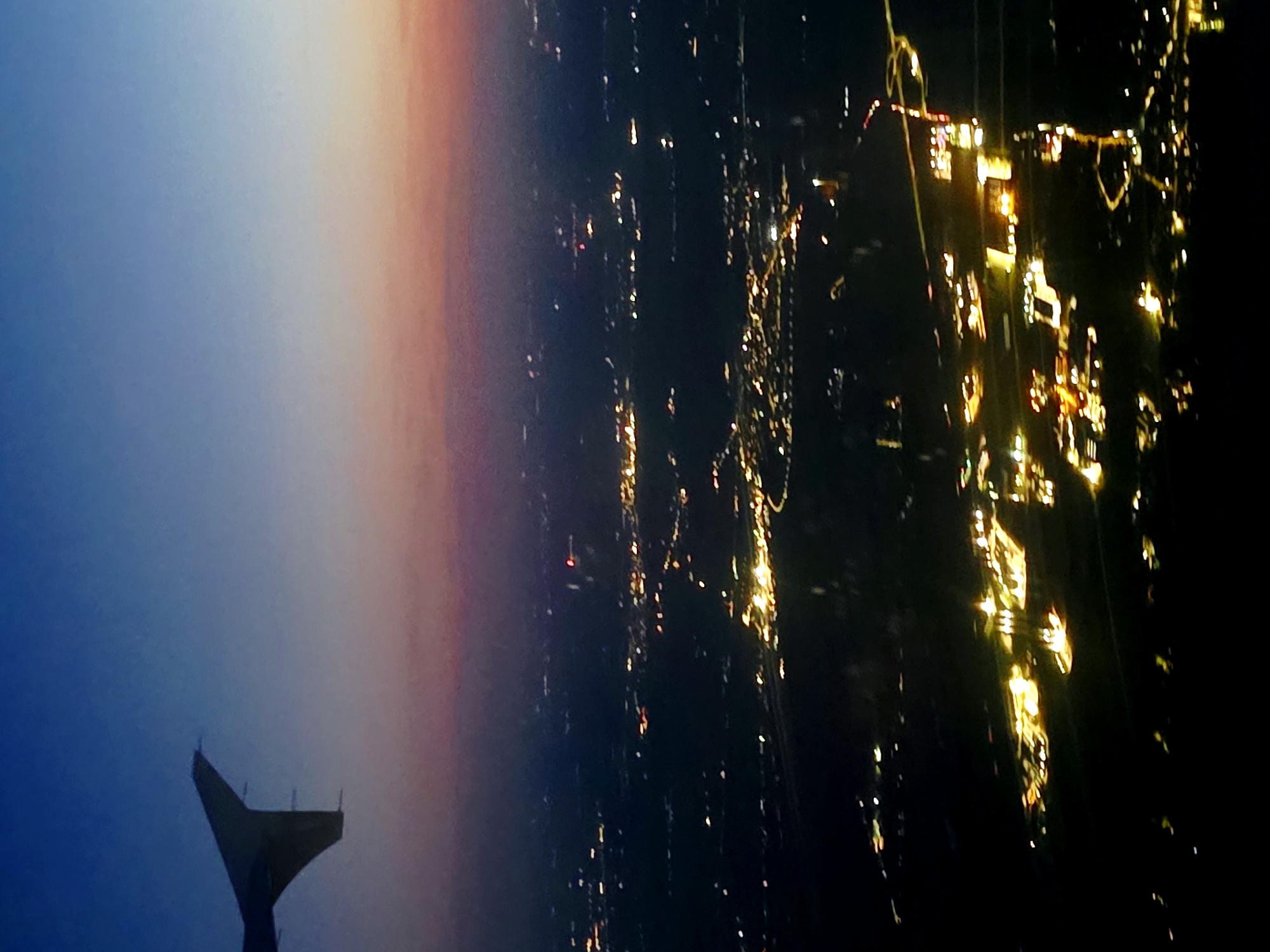 The image is an aerial view of the city of Pokrzywnica, Poland, taken at dusk. The sky is a gradient of deep blue at the top, fading into purple and then a soft orange-pink near the horizon. A portion of an airplane wing is visible in the upper left corner, silhouetted against the sky. Below the sky, the city is illuminated by numerous lights, suggesting a developed area with buildings, roads, and possibly industrial sites. The lights create a speckled pattern across the dark landscape, indicating the presence of human activity and infrastructure. The overall scene is peaceful and serene, capturing the transition from day to night over an urban environment. The specific location within Pokrzywnica is not discernible from the visual information provided.