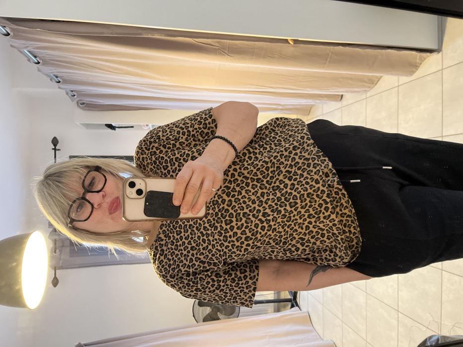 In the city of Chambœuf, France, an indoor scene depicts a middle-aged woman taking a mirror selfie. The woman, with blonde hair and black-rimmed glasses, is wearing a brown and black leopard-print short-sleeved top and black sweatpants with a visible drawstring. Her left hand is partially in her pocket, revealing a dark tattoo on her forearm and a silver ring on her left ring finger. A dark beaded bracelet is on her right wrist. She holds a light-colored smartphone with a dark rectangular grip on its back in her right hand, extended to capture her reflection. Her facial expression is playful, with her tongue slightly extended to the left. The background, reflected in the mirror, shows a light-colored tiled floor and walls. An overhead light fixture is visible at the top. To the left, a standing fan and a drawn light-colored curtain are present. To the right, a series of light-colored curtains with silver grommets suggests an adjoining space, possibly a dressing area. A white shelf is visible in the upper right background.