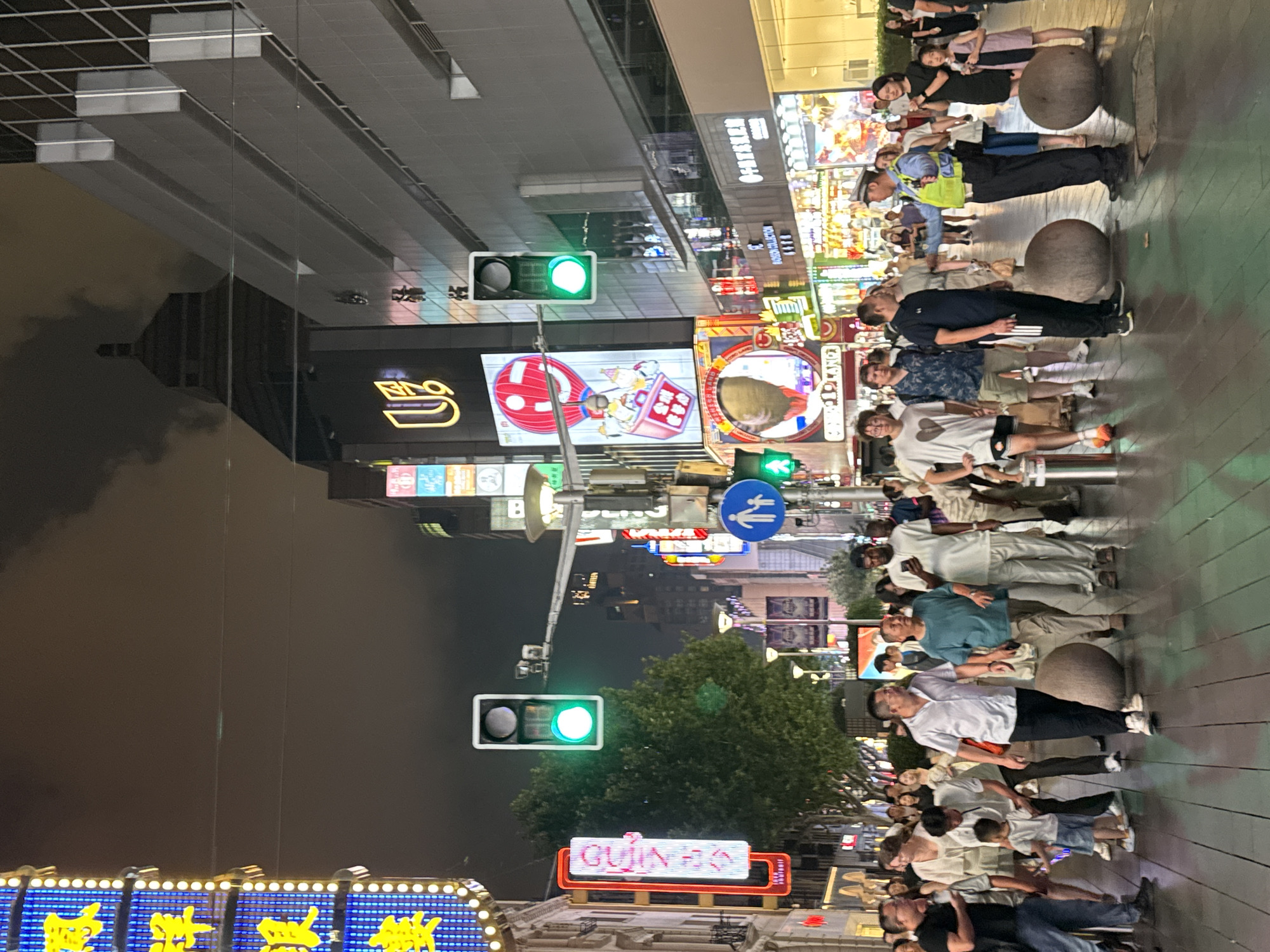 The image depicts a bustling street scene at night in Shanghai, China, likely in a commercial or entertainment district. The environment is illuminated by numerous bright neon signs and digital billboards displaying advertisements in Chinese characters and graphics. A traffic light hangs overhead, showing a green signal. A diverse group of pedestrians, including men, women, and children, are walking on a paved pedestrian walkway. Some individuals appear to be conversing, while others are simply moving through the area. A police officer in uniform is visible interacting with a civilian. The architecture in the background features tall modern buildings with glass facades and traditional Chinese-inspired roof elements. The overall atmosphere suggests a lively urban environment at night.