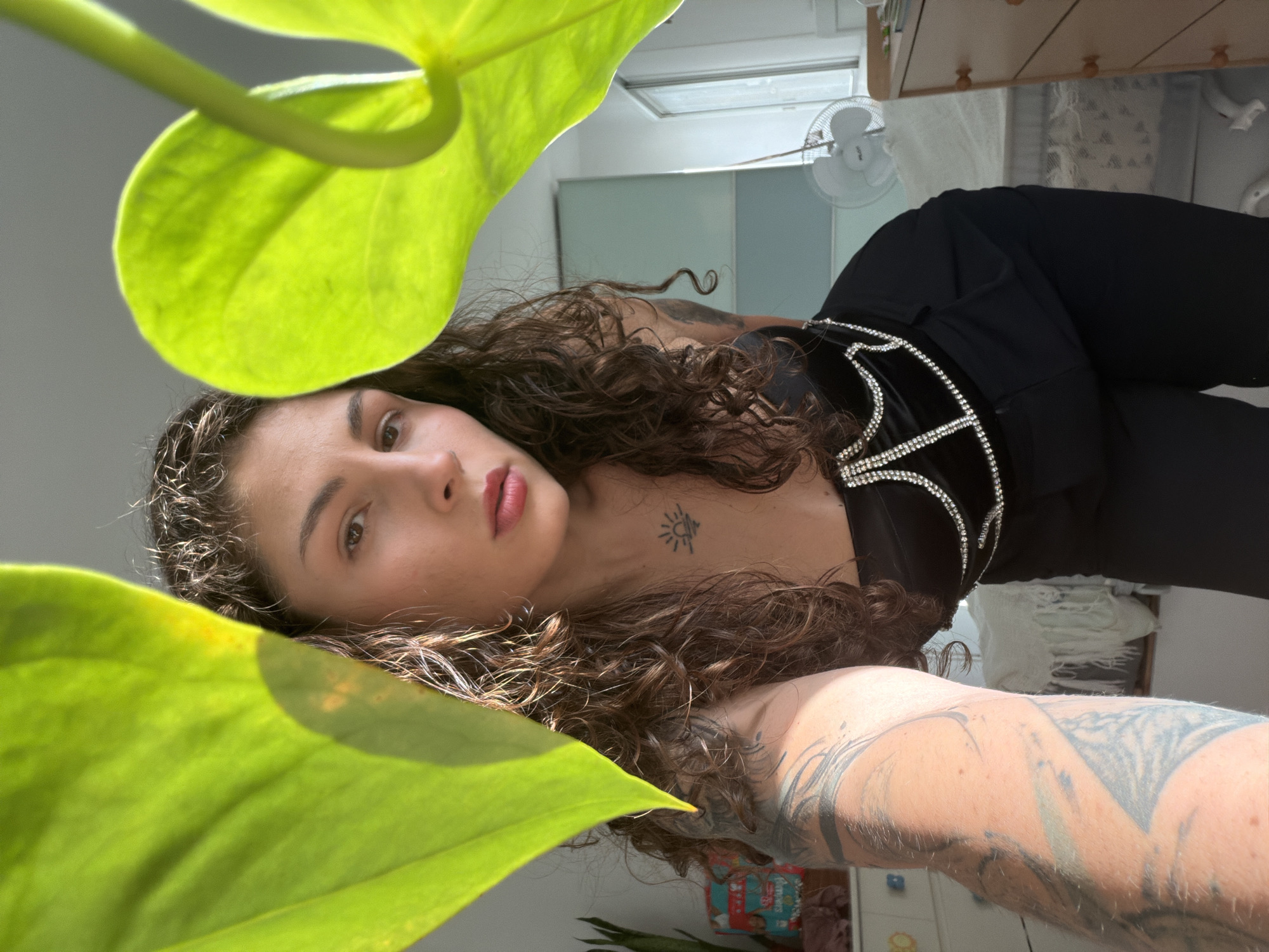 A woman with curly brown hair is posing with a large, green leaf in the foreground. She is wearing a black top with diamond-like embellishments and black pants. The woman has tattoos on her chest and arm. Behind her is a room with a fan, window, and dresser. The location is likely an indoor space, potentially in a home or apartment, somewhere in Israel. Kiryat Bialik's potential relevance is unknown as no clear markers confirming the location are visible.