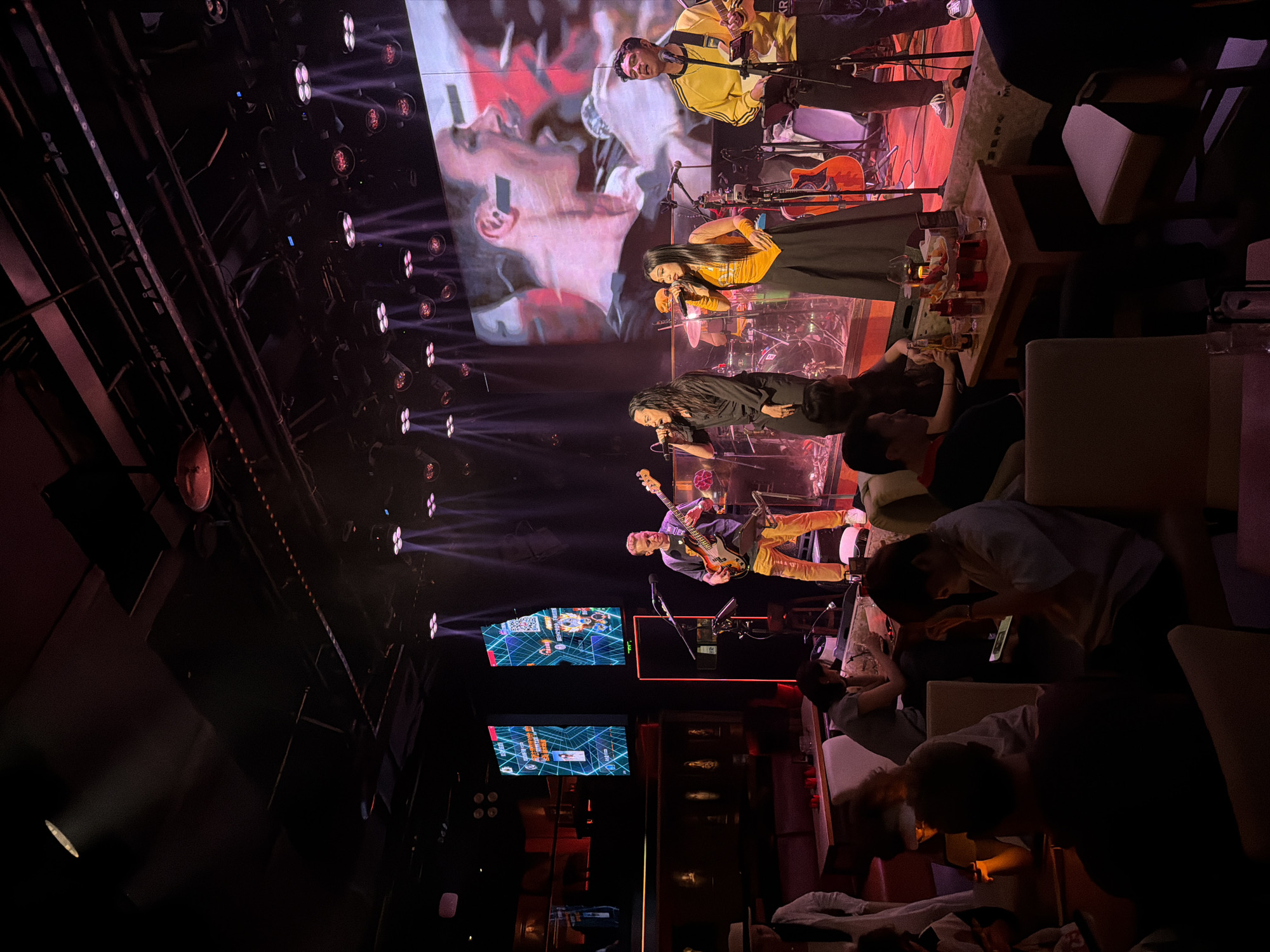 The image shows a band performing on a stage in a dimly lit venue. The band consists of four members: a male bassist in the foreground on the left, a male singer in the middle left, a female singer in the middle right, and a male guitarist on the right. They are all illuminated by stage lights. Behind them, a large screen displays a close-up of a person's face. The venue appears to be a club or a bar, with tables and seating in the foreground where audience members are seated, some of whom are looking at their phones. The overall atmosphere is that of a live music performance.