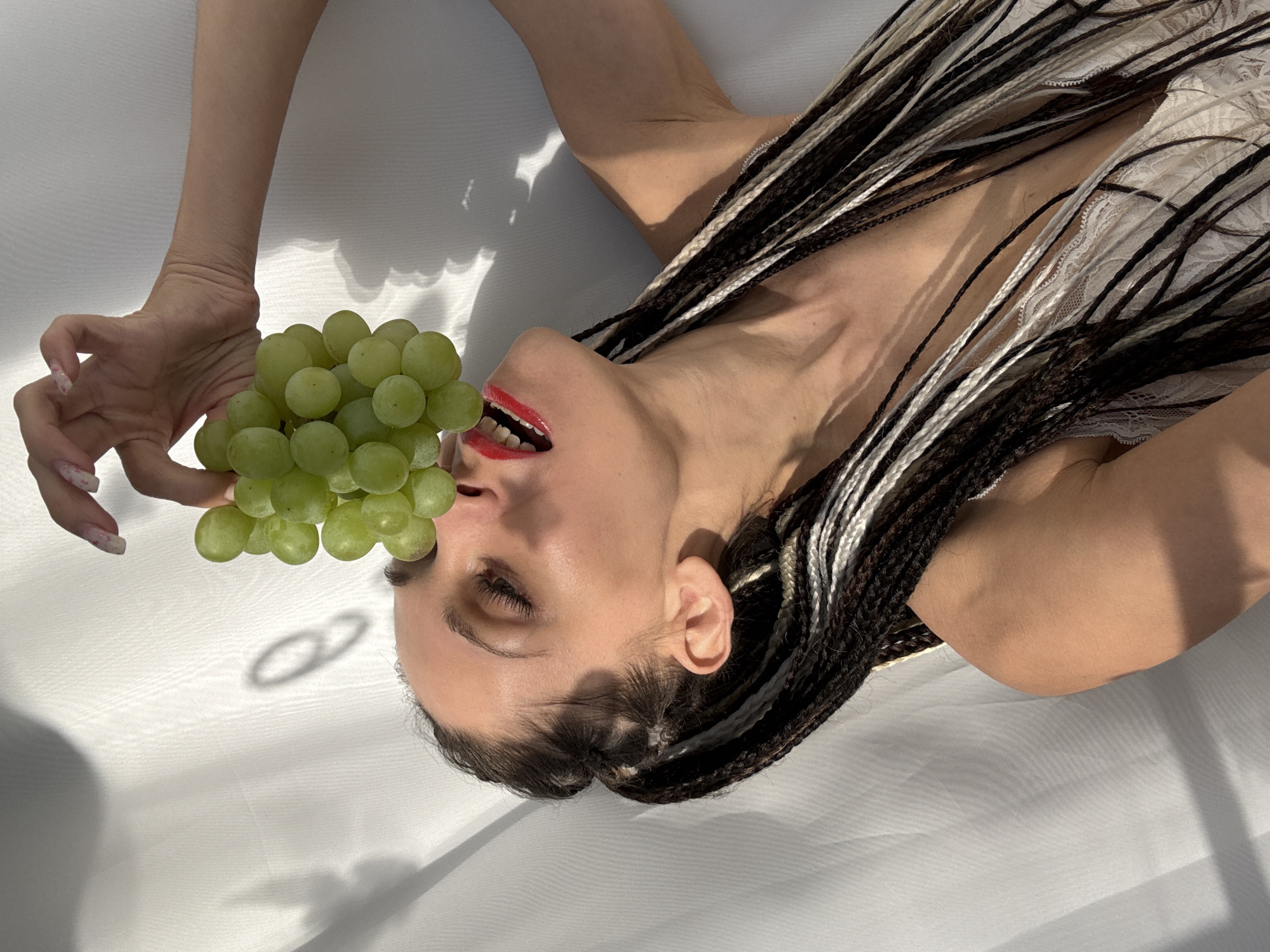 A person with braids and red lipstick lies on a white surface, holding a bunch of green grapes near their mouth. The location is not discernible from the image and thus Rusanovo, Russia cannot be confirmed.