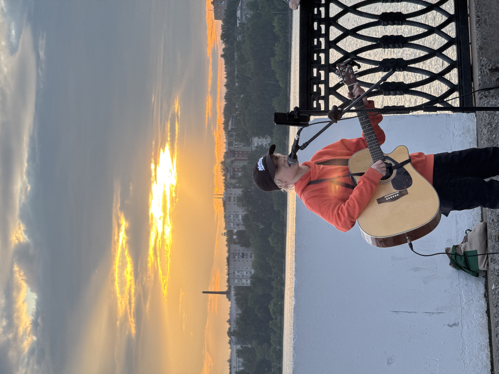 The image depicts a young person playing an acoustic guitar and singing into a microphone outdoors, with a body of water and a cityscape in the background. The setting appears to be a promenade or embankment, suggested by the white concrete wall on the left and the decorative black metal railing on the right. The cityscape features apartment buildings and a tall industrial smokestack, characteristic of many Russian cities. The sky is filled with dramatic sunset clouds, with warm orange and yellow hues illuminating the scene.
The individual is wearing an orange hooded sweatshirt and a black baseball cap. They are holding a light-colored acoustic guitar and their hands are positioned as if playing. A microphone is set up on a stand in front of them. On the ground near the person is a beige bag with green accents, to which a black cable from the guitar is connected. The body of water reflects the warm light of the sunset. The overall atmosphere is one of performance or busking.
This scene is highly likely to be located in Izhevsk, Russia, given the presence of typical Soviet-era apartment blocks and an industrial smokestack, which are common architectural features in many Russian cities, including Izhevsk. The specific visual elements align with waterfront areas in Izhevsk that are often used for recreation and public gatherings.