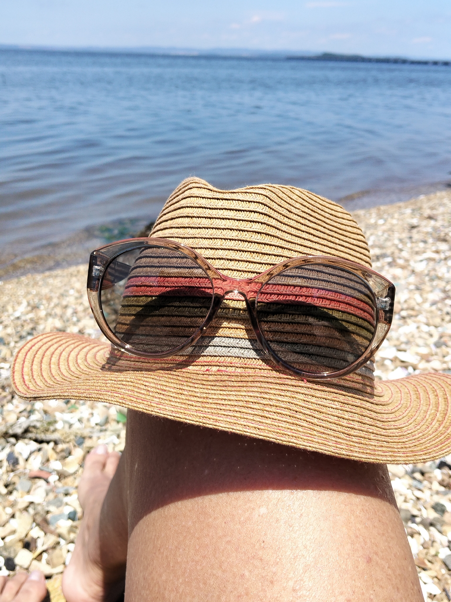 The image shows a close-up of a straw hat with sunglasses resting on it, placed on a person's leg. The hat has alternating bands of tan and lighter straw, with subtle red stripes. The sunglasses have clear frames and dark lenses, reflecting the hat's pattern. In the background, the sea is visible, with a shoreline of pebbles and sand. The sky is a clear blue. The overall scene suggests a relaxed day at the beach. The image is taken from a low angle, focusing on the hat and the person's leg, with the water and sky forming the backdrop. There is no indication of this location being in Vladivostok, Russia.
