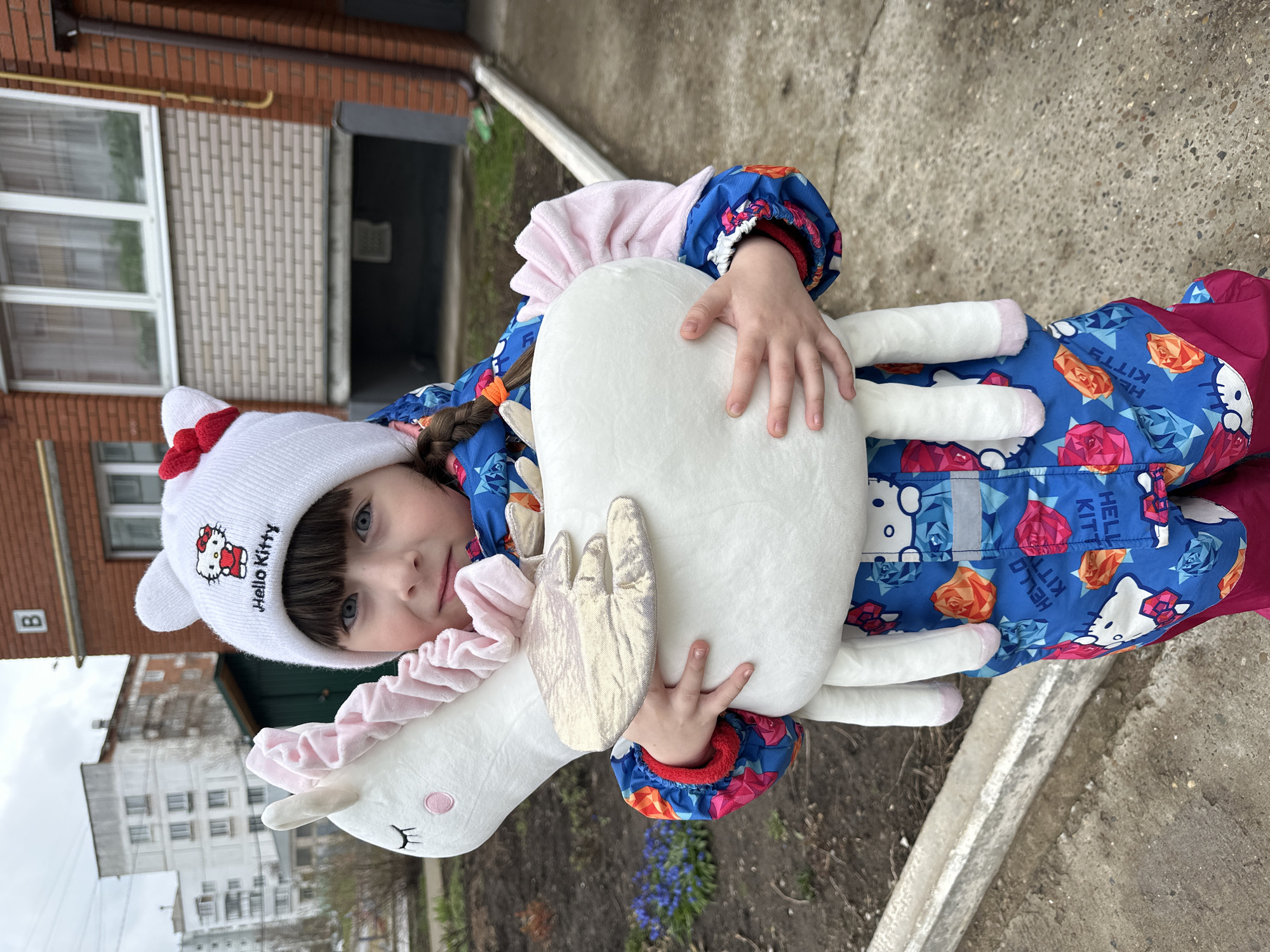 A young child, likely a girl, stands outdoors in Chaikovsky, Russia, holding a large white plush unicorn. The child, with dark hair, bangs, and blue eyes, looks directly at the camera with a neutral expression. She wears a white knitted beanie hat adorned with a red bow and the embroidered text "Hello Kitty." Her outerwear is a vibrant blue and pink winter coat or overall, patterned with "Hello Kitty" faces and geometric rose shapes, also featuring the text "Hello Kitty."
The plush unicorn has a pink ruffled mane, small golden wings, and subtle pink blush on its cheek. The background features urban elements: a brick building with white-framed windows, a dark doorway, and patterned siding. Another, more distant building with multiple windows is visible. The ground is paved, with a concrete curb and patches of dry earth where a few blue flowers (possibly Muscari) are beginning to bloom. The lighting is soft and diffused, suggesting an overcast day or early spring conditions, consistent with the child's warm attire. The letter "B" is visible on a sign on the brick building. The image captures a casual moment outdoors.