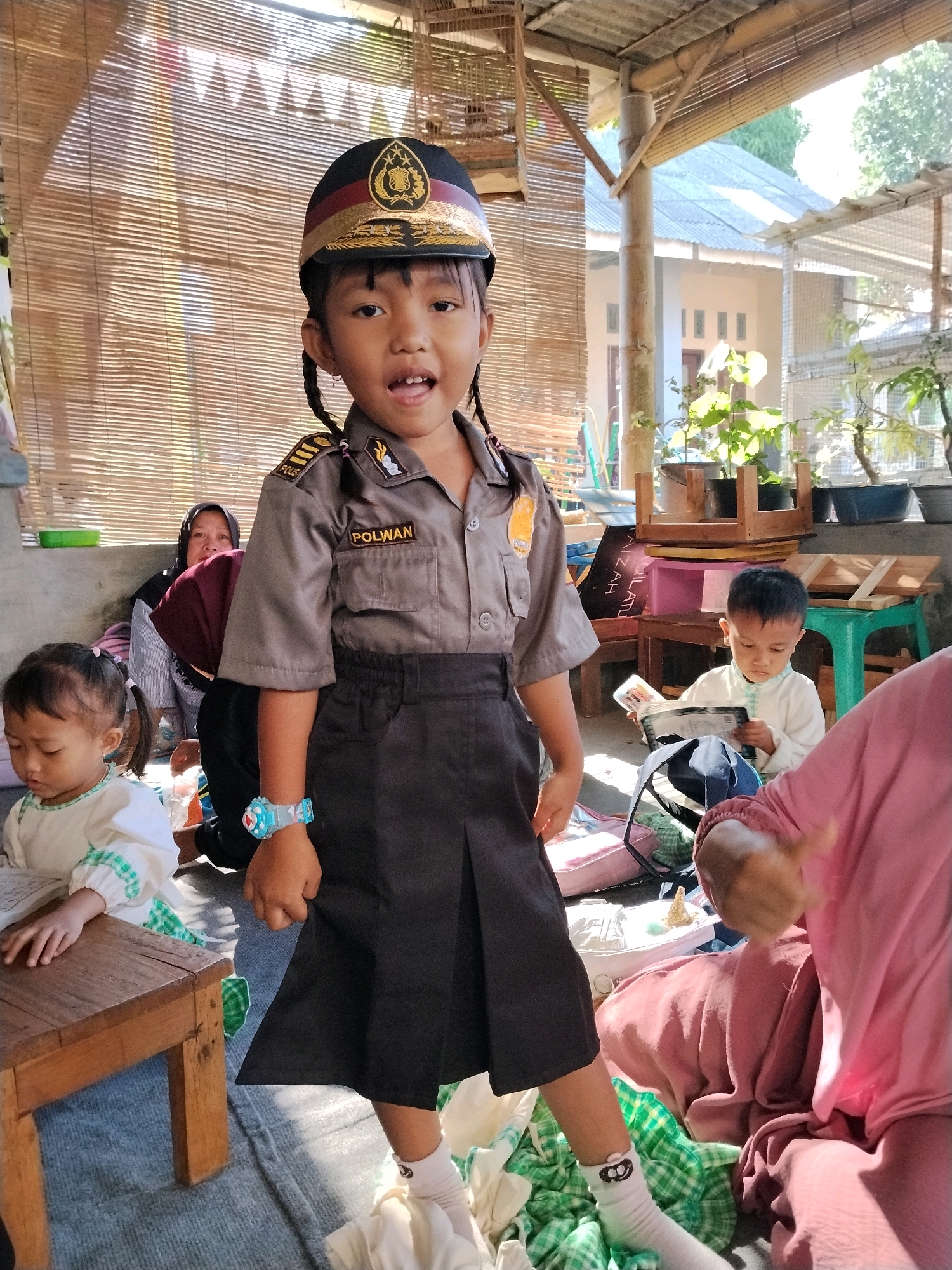 In Panggungan, Indonesia, a young Indonesian girl stands in the foreground, dressed in a police uniform. She wears a dark cap with gold trim and an emblem, a gray shirt with "POLWAN" written on the chest and epaulets, and a dark pleated skirt. A colorful watch adorns her left wrist. Her hair is braided in two pigtails.
Behind her, a woman in a headscarf and maroon hijab sits with her back to the camera, partially obscured by the girl. To the left of the girl, a younger child in a white outfit with green trim is seated, looking down. Further in the background, another child in a white shirt is reading a book. The scene appears to be outdoors, with bamboo blinds and corrugated metal roofing visible, suggesting a simple, open-air structure. Potted plants are present, and sunlight casts shadows, indicating daytime. The overall setting is informal and appears to be a gathering of children and adults.