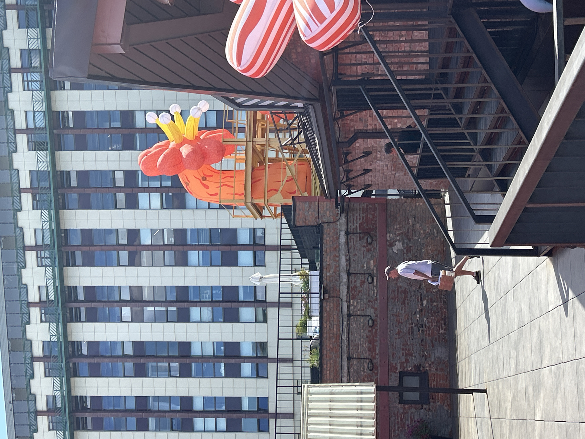 An outdoor scene, likely a rooftop or elevated patio, features several elements. In the background, a tall, modern building with a grid of windows is visible. In the mid-ground, a large, orange inflatable dragon-like structure is prominent, positioned on a yellow scaffolding. To the right, a dark-roofed building with brick accents and an external staircase is present. In the foreground, a man in a white shirt and grey shorts walks across a tiled patio, carrying a cardboard box. Another person is partially visible on the staircase of the brick building. Candy cane-shaped inflatables are also present, hanging near the roof of the brick structure.