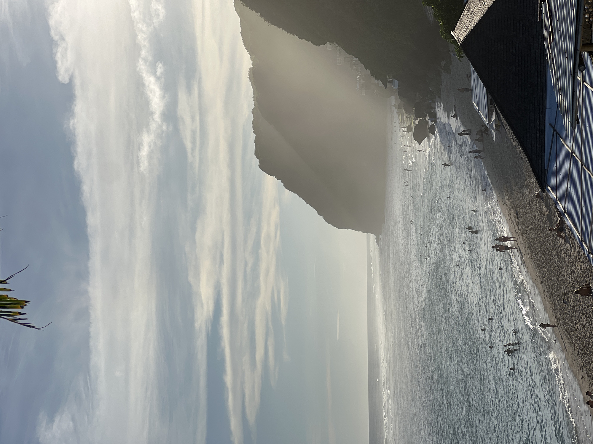 This image captures a vibrant beach scene, likely in Kuta Selatan, Indonesia, during what appears to be late afternoon or early evening due to the lighting. A majestic, tree-covered mountain or cliff dominates the left side of the frame, receding into a hazy distance, suggesting atmospheric perspective or backlighting from the sun. Along the mountain's base, some faint structures are visible within the haze.
The central and right portions of the image feature the ocean, its surface shimmering with gentle waves reflecting the bright sky. Numerous people are scattered across the water, swimming, wading, and enjoying the ocean. Closer to the foreground, a dark sandy or pebbly beach is visible, where several individuals walk along the shoreline or stand near the water's edge.
On the far right, buildings with dark roofs and textured facades, possibly beachfront hotels or establishments, line the coast. The sky above is a pale blue, streaked with long, wispy white clouds. A few dark green palm fronds are visible in the extreme top left corner, contributing to the tropical setting. The overall atmosphere is one of leisure and relaxation, with many people engaging in beach activities under a bright, hazy sky. There is no visible text in the image.