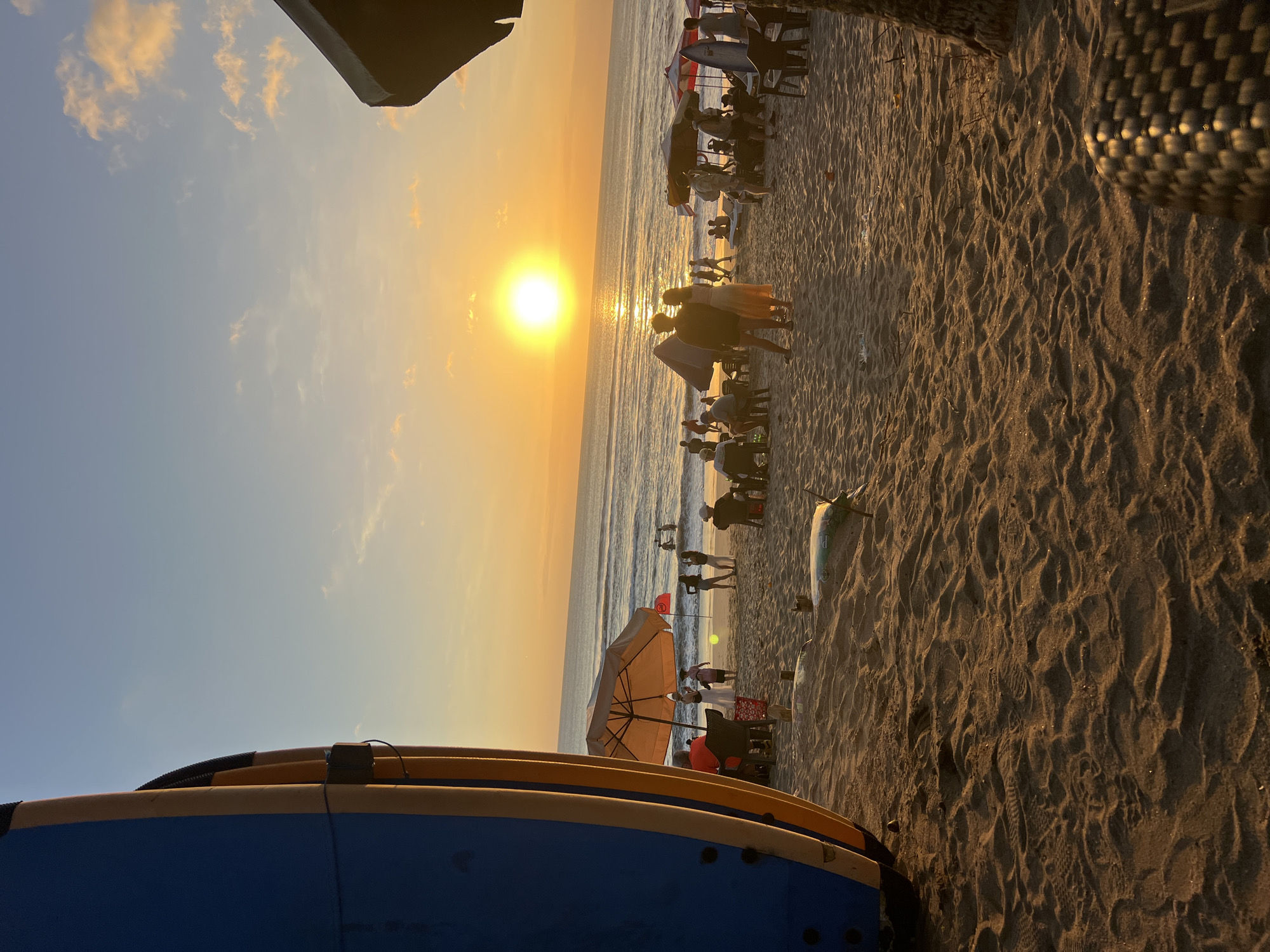 The image depicts a sunset scene on Kuta Beach, Indonesia. The sun, a bright orb, is low on the horizon, casting a warm golden glow across the sky and the ocean. Waves are gently rolling onto the sandy shore.
In the foreground, the left side of the frame is dominated by the blue and yellow side of several surfboards stacked upright. The sand in the foreground is textured with ripples and footprints.
Further back, people are scattered across the beach. Some are seated under beach umbrellas, possibly at a cafe or restaurant. Others are standing or walking. A couple stands close together near the water's edge. In the ocean, a few figures can be seen swimming or surfing. Beach umbrellas, chairs, and tables are visible, suggesting a lively beach scene. The overall atmosphere is tranquil and picturesque, characteristic of a tropical beach sunset.