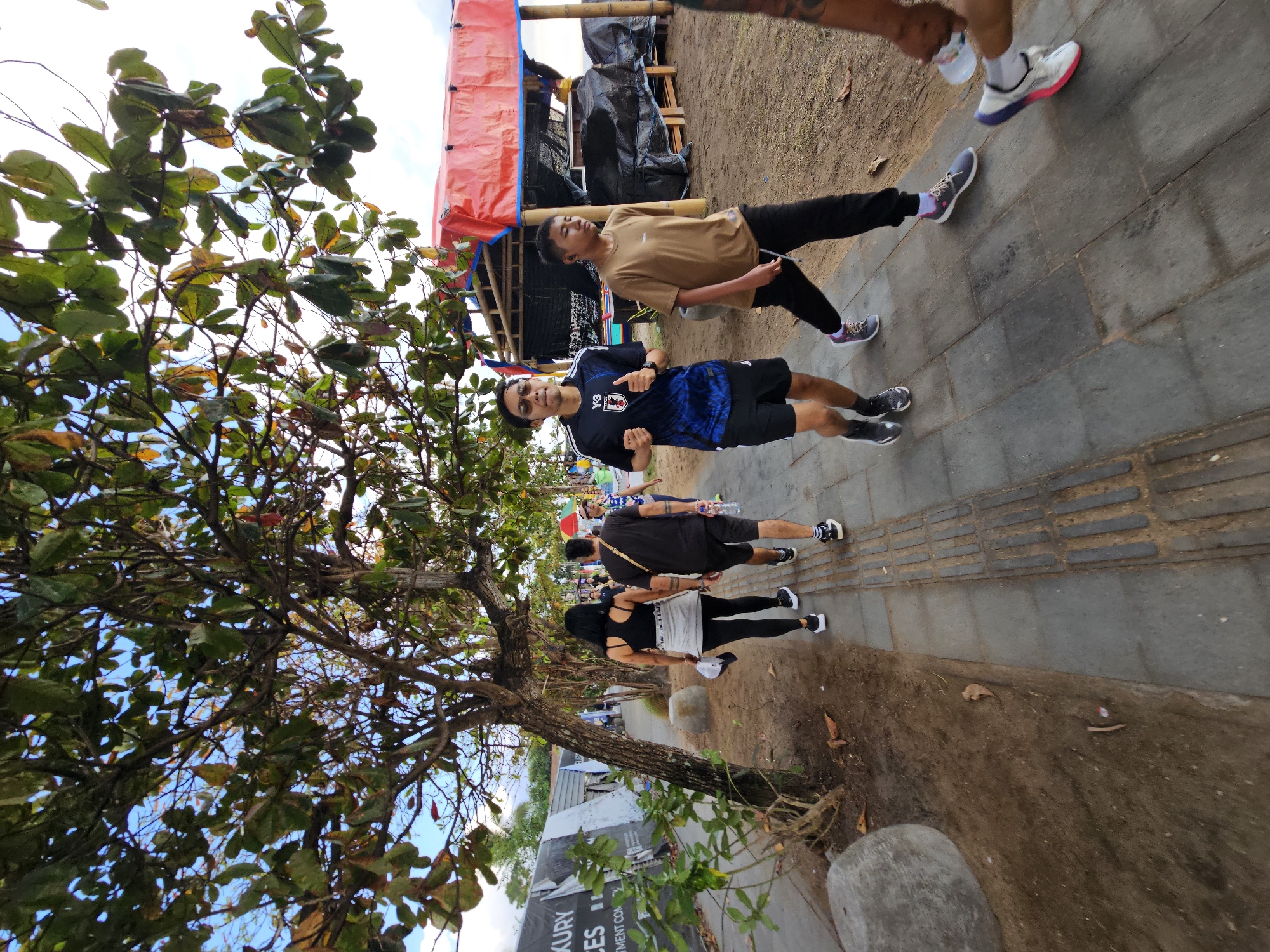 The image depicts a street scene in Denpasar, Indonesia, likely near a beach or coastal area, given the sandy ground and tropical foliage. In the foreground, a group of people are walking and running along a paved walkway. The individuals include a man in a blue and black athletic shirt and black shorts, running with his thumbs up. To his right, a younger person in a tan t-shirt and black pants is walking. Behind them, a woman in a black tank top and grey skirt and a man in a black t-shirt and black shorts are walking together, with the woman holding a white cap. Further back, another person is visible. The environment features a large tree with green leaves and branches casting shadows on the walkway. In the background, there are temporary structures covered with red tarps and bamboo poles, suggesting a market or roadside vendors. A black tarp is visible behind the red coverings. The sky is partly cloudy with a bright light source. The overall impression is a casual, outdoor activity scene in an urban or semi-urban coastal setting.