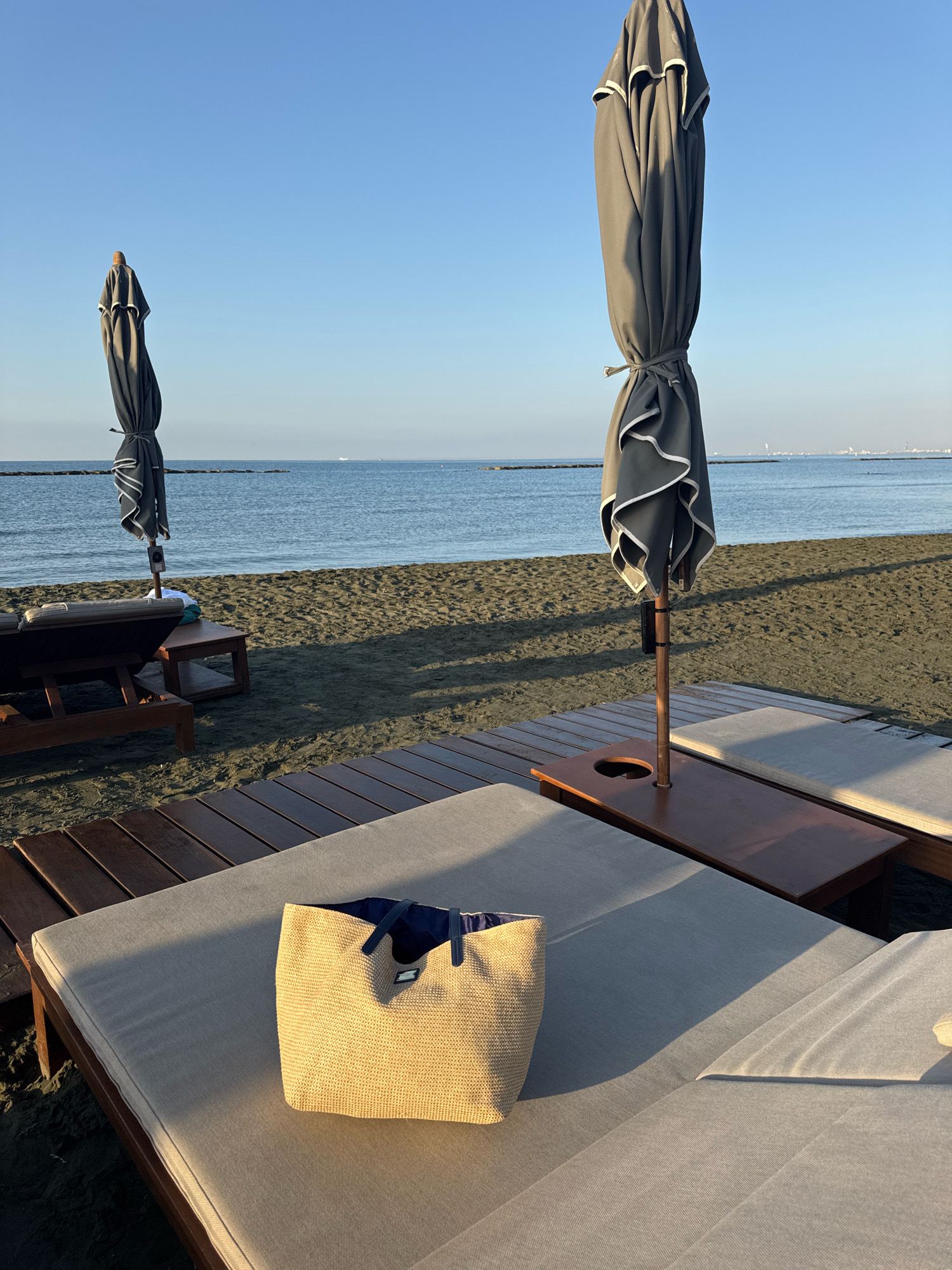 This image depicts a serene beach scene in Agios Tychona Community, Cyprus, bathed in the warm glow of morning or late afternoon sun. In the foreground, a light-colored woven straw tote bag rests on a cushioned lounge chair. The chair itself is part of a set, with another similar chair and a wooden side table visible. Beyond the lounge chairs, a dark wooden deck extends towards the sandy beach, which is a mixture of dark sand and small pebbles.
Two large, dark grey beach umbrellas, folded and tied, stand sentinel on the wooden deck. The ocean stretches out beyond the sand, its surface calm and reflecting the clear, pale blue sky. A distant line of breakwaters or a shallow reef runs parallel to the coastline. In the far distance, a faint outline of a city or industrial area can be seen on the horizon. The overall atmosphere is one of tranquility and peacefulness, suggesting an undisturbed moment at a beach resort. There are no people visible in the image.