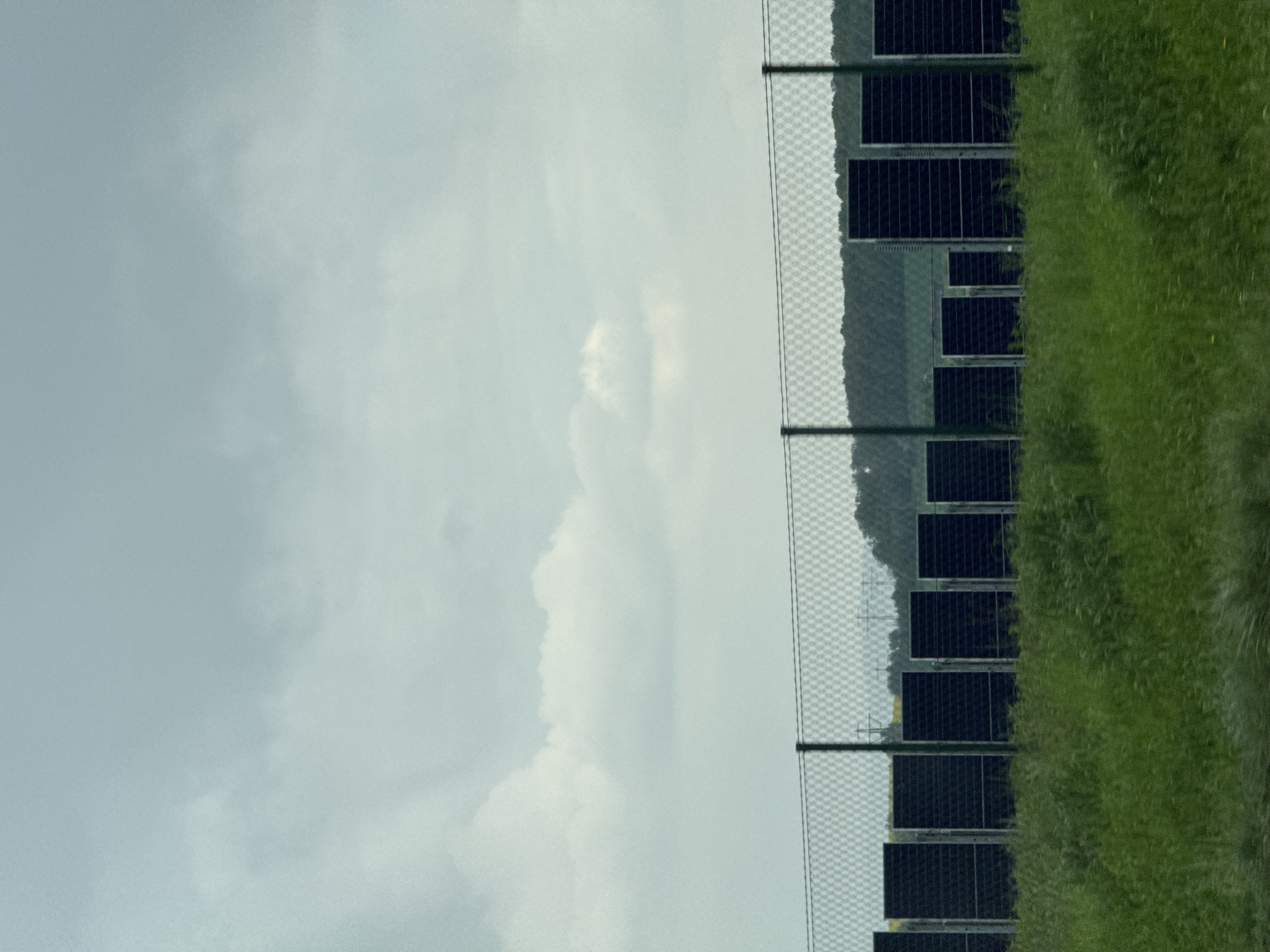 This image captures a landscape in Bad Rappenau, Germany, featuring a solar farm under an overcast sky. The upper half of the frame is dominated by a light grey to off-white cloudy sky, indicating diffused lighting conditions.
A wire mesh fence, with dark vertical support posts, runs horizontally across the mid-ground, acting as a boundary. Behind this fence, numerous dark, rectangular solar panels are arranged in rows, signifying a solar power generation facility. In the far background, beyond the solar panels, a line of dark green hills or dense tree lines forms the distant horizon. Faintly visible in the background, among the hills, are structures resembling utility poles or power lines.
To the right side of the frame, occupying the foreground, is a dense patch of lush, tall green grass. No specific activity or event is taking place, and no people are visible. The time of day appears to be either morning or afternoon on a cloudy day, given the soft, uniform light. There is no discernible text within the image. The scene presents a blend of modern renewable energy infrastructure with a natural, tranquil environment.