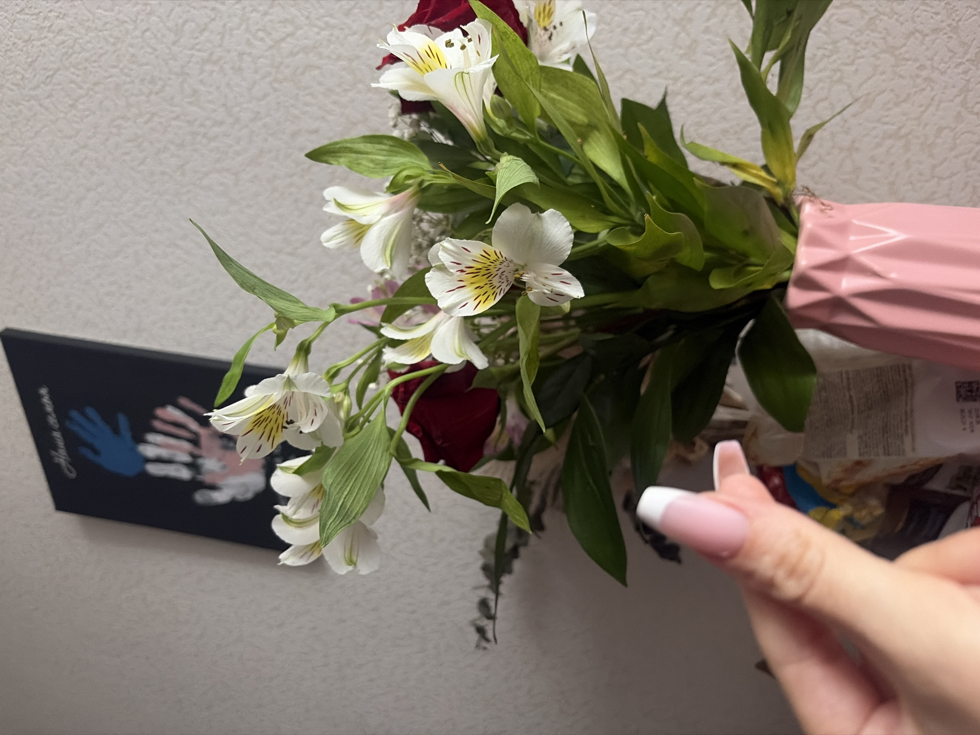 A medium-angle shot shows a bouquet of flowers, primarily white Alstroemeria and some red roses, in a pink vase with a geometric pattern. The flowers and vase are positioned against a textured, light gray wall. To the left, a black rectangular canvas with handprints in blue, white, and pink is visible. In the lower right foreground, a person's hand with a pink manicure and white tips is partially visible. The location is likely an indoor setting, but there are no indicators suggesting the location is Saint Petersburg, Russia.