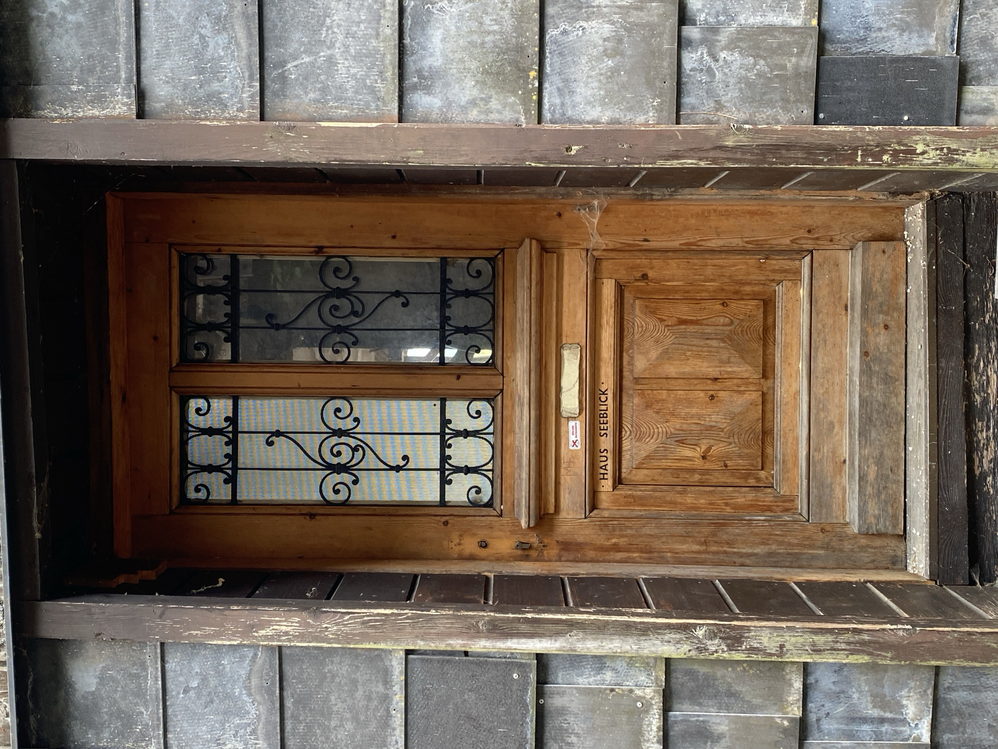 The image shows a close-up view of a weathered wooden door in Hallstatt, Austria. The door is made of light brown wood and features two glass panes with ornate black wrought-iron grilles at the top. Below the glass panes, there is a mail slot and a small red sticker with white text. Centered on the lower part of the door, there is carved text that reads "HAUS SEEBLICK". The door is set within a dark wooden frame, which is flanked by grey, slate-like siding. The entrance also includes wooden steps leading up to the door. The overall scene suggests an older, rustic building.