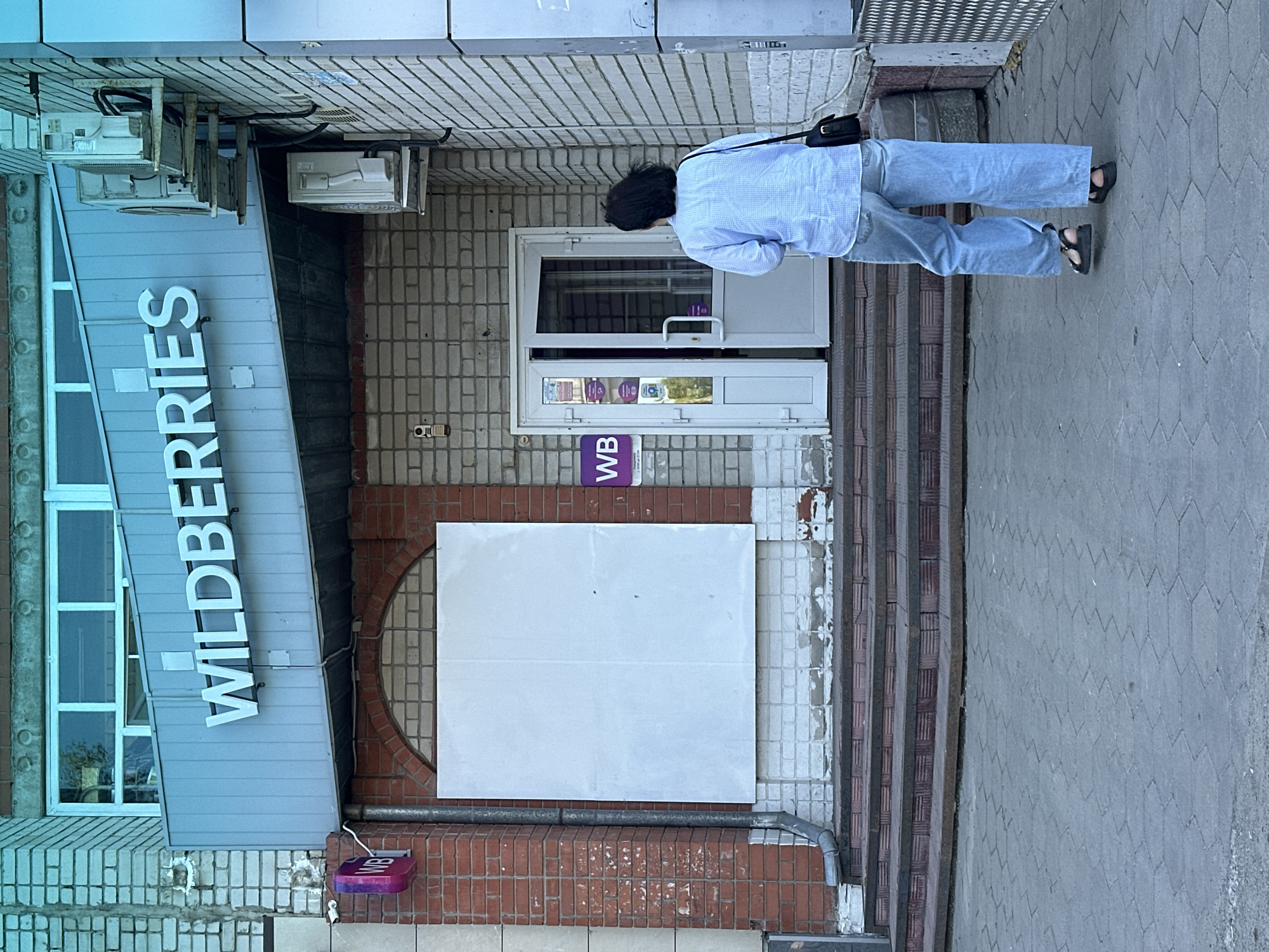 An exterior shot of a Wildberries store entrance is captured from a low angle, showcasing a person from behind as they approach the doorway. The store's prominent "WILDERRIES" sign is mounted above a light-colored awning, contrasting with the brick facade of the building. To the left of the entrance, a blank, white square panel is affixed to the brickwork, possibly a former advertisement space. The entrance itself features double glass doors, one of which is partially ajar, revealing a glimpse of the interior. A purple "WB" logo is visible to the left of the doorway. Two air conditioning units are mounted on the brick wall to the right of the store sign. The ground in front of the store is paved with hexagonal gray tiles, and a set of tiled stairs leads up to the entrance. The overall environment suggests a commercial street in the city of Novoaltaysk, Russia.