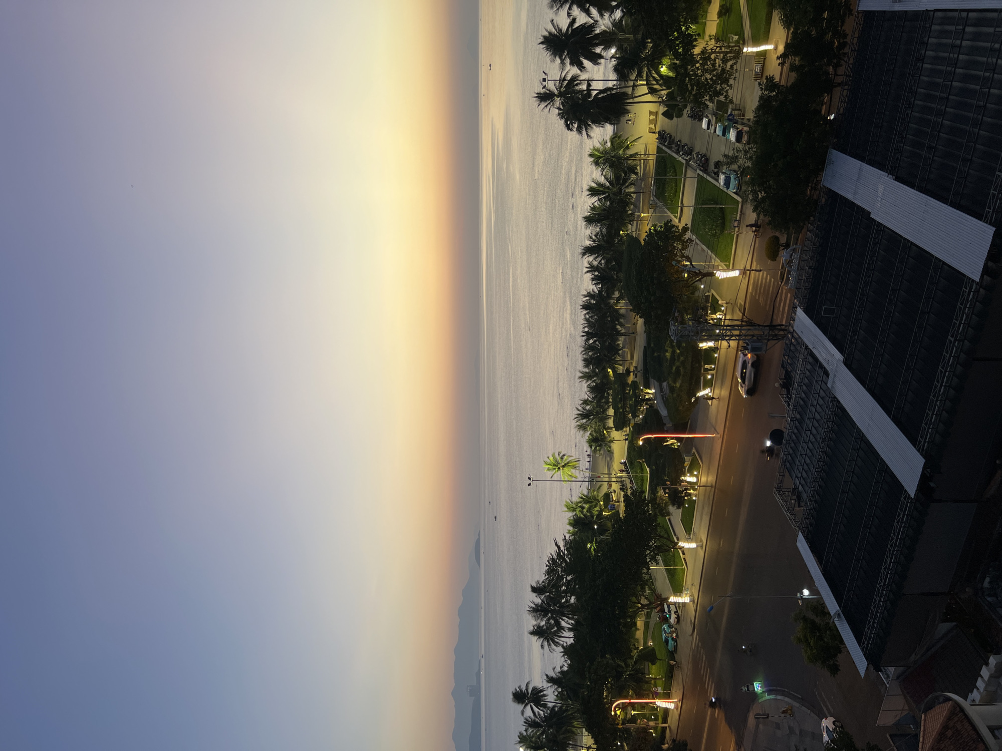 This image captures a coastal view of Nha Trang, Vietnam, during twilight. The foreground features the roof of a building with a corrugated metal surface. Beyond this, a street runs parallel to the coastline, with vehicles including cars and motorcycles visible. A pedestrian crossing is marked on the road.
Adjacent to the street is a park-like area with palm trees, well-maintained grassy patches, and decorative lighting. Several streetlights illuminate the scene. Further back, the sandy beach is visible, dotted with people and more palm trees. The sea stretches out towards the horizon, where a line of distant mountains or hills can be seen. The sky displays a gradient of colors characteristic of sunset or sunrise, transitioning from a pale yellow near the horizon to shades of blue at the top. A few boats are visible on the calm water.