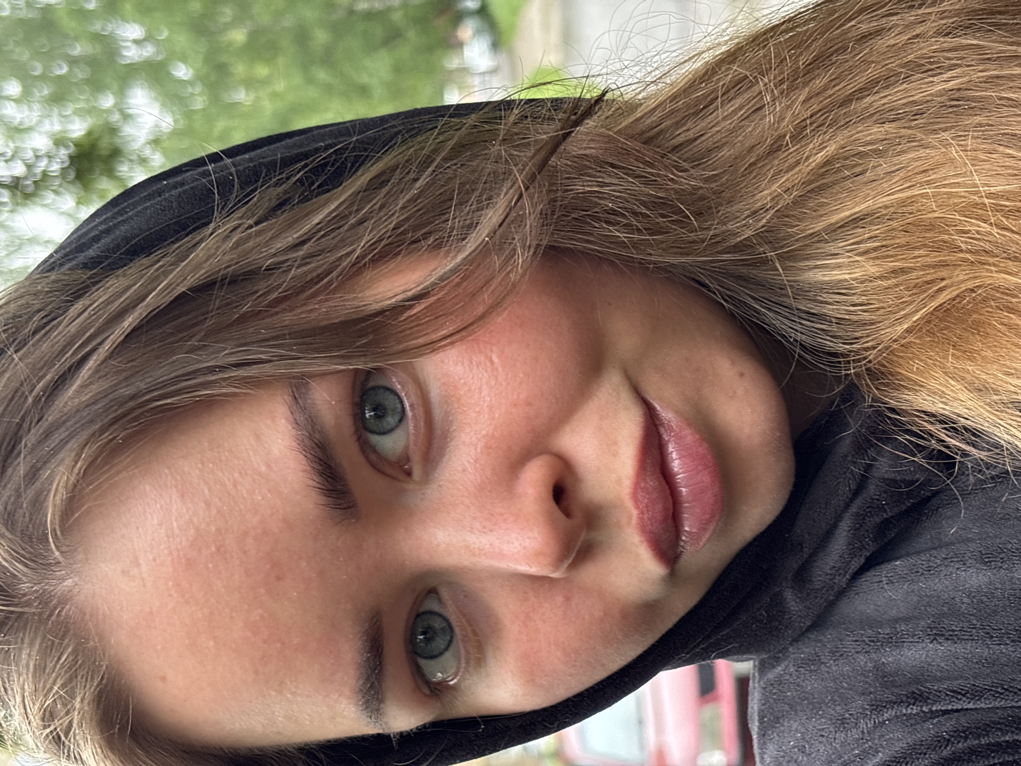 A young woman with light skin, light brown hair, and blue-green eyes is wearing a black hooded garment. She appears to be outdoors, possibly in a park or residential area. The background is blurred, showing green foliage and indistinct shapes, and her head is tilted slightly. No specific details tie this image to the city of Izhevsk, Russia.
