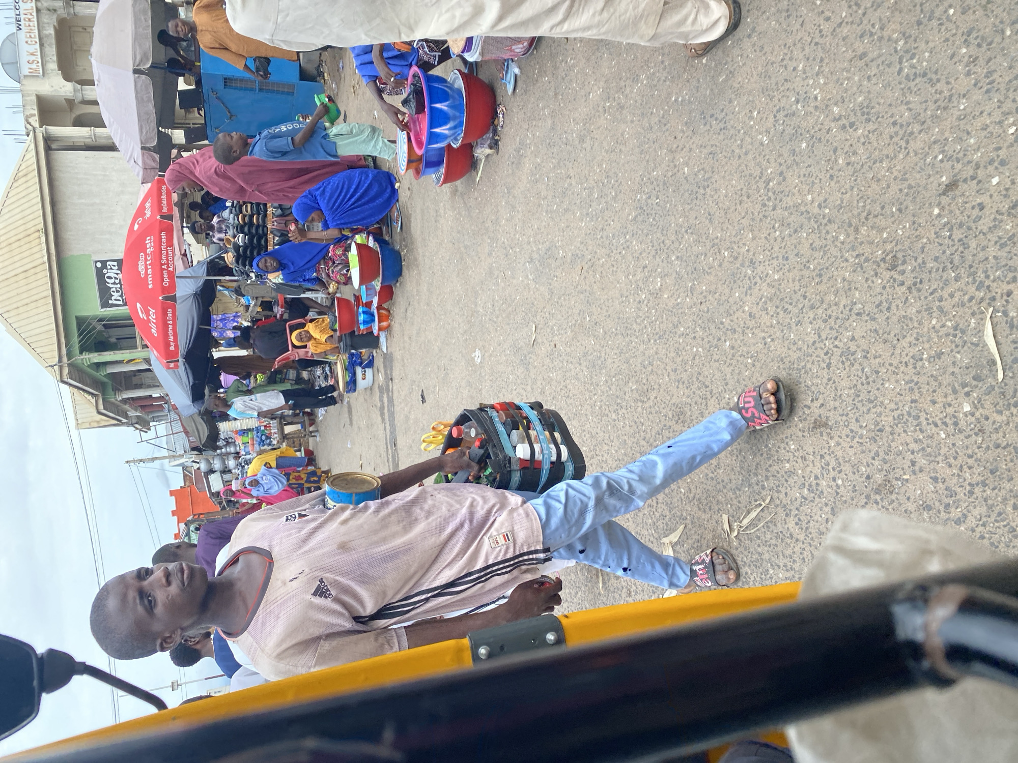 This image captures a bustling street scene in Jalingo, Nigeria, likely during daylight hours given the bright lighting and shadows. The primary subjects are several people engaged in commerce at a market. In the foreground, a young boy, wearing a light-colored, striped t-shirt and light blue jeans, is the most prominent figure. He is looking towards the camera with a neutral expression. He appears to be on or near a yellow three-wheeled vehicle, possibly a motorcycle rickshaw, suggested by the yellow bar in the foreground and the vehicle's structure.
Behind the boy, other individuals are visible, some interacting with goods and others standing or walking. Several women are seen wearing colorful headscarves, a common attire in Nigeria. One woman, in a bright blue headscarf and dress, is surrounded by an assortment of plastic bowls in various colors, including red and blue. Another person, dressed in a maroon garment and headscarf, seems to be attending to the market stall.
The market appears to be set up along a paved street, with several stalls and shops in the background. An umbrella with the "bet9ja" logo is visible, suggesting a commercial area with various services. There are also numerous items for sale, including what appear to be more plastic bowls, containers, and potentially food items or other wares arranged on shelves and tables. The overall atmosphere seems active and typical of a vibrant marketplace. The ground is a mix of asphalt and gravel, with some scattered debris, indicating a busy public space.