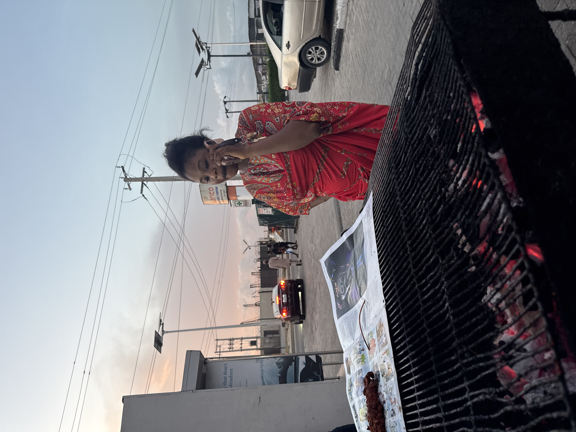 A woman in a red floral dress is talking on a pink cell phone, standing on a sidewalk in Ado, Nigeria. In the foreground, a newspaper is laid out on a table beside a barbecue grill filled with glowing coals. Skewered meat rests on the newspaper. Behind the woman is a street with a parked silver car and a red car driving away. Pedestrians are visible in the distance. Utility poles and lines are present. A building with a sign that reads "ENCO CENTRE" is visible, as is a sign that reads "days that don't calendars". The sky is a hazy orange-pink.