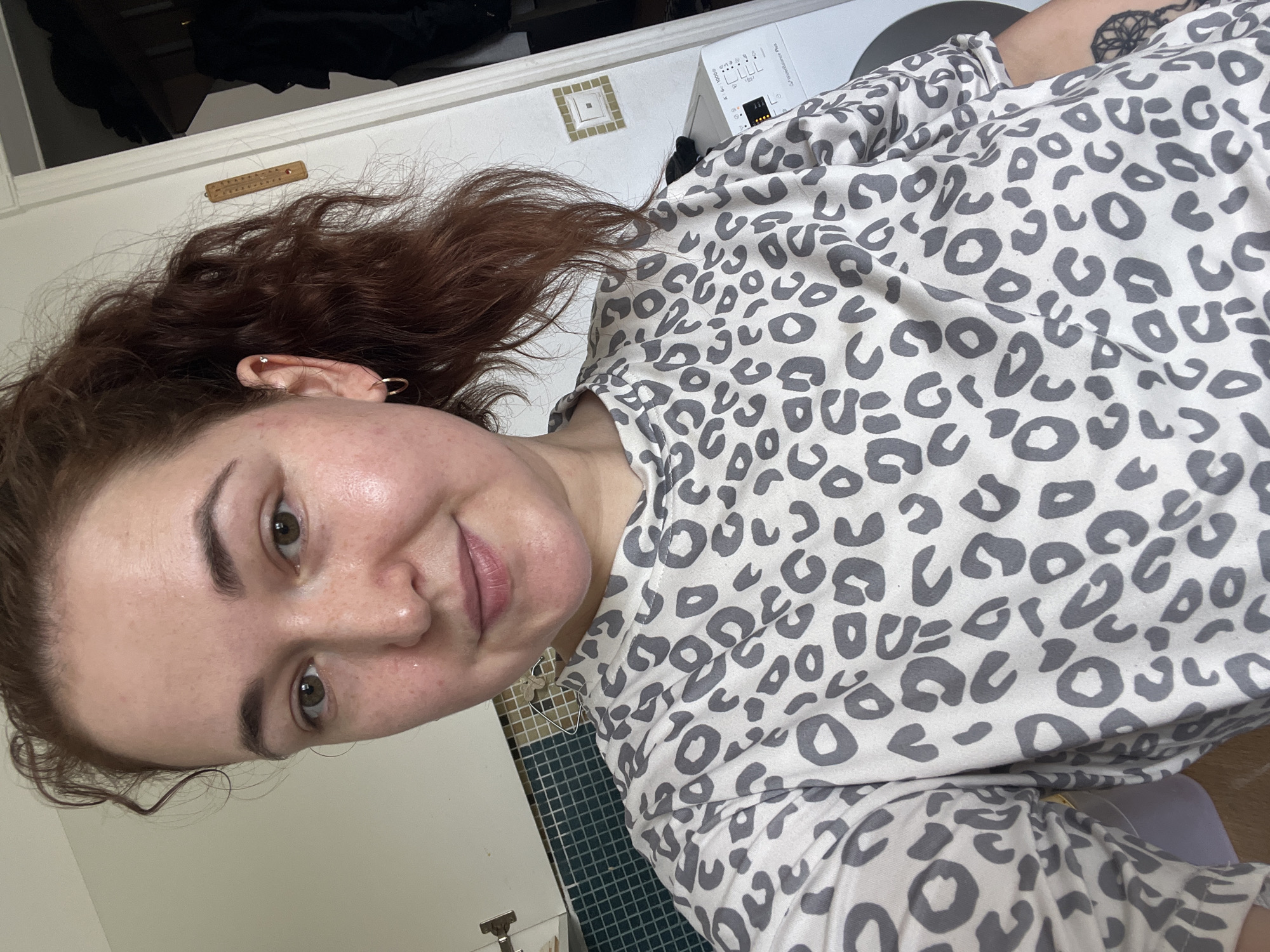 A Caucasian woman with reddish-brown hair pulled into a ponytail is shown from the chest up, looking at the camera. She is wearing a light-colored shirt with a gray leopard print pattern. Her skin appears slightly shiny, and she has freckles on her face. Her right ear has two piercings, one with a small stud and another with a hoop earring. Behind her, to the left, is a white tiled backsplash in a kitchen area, with a white cabinet door visible. To the right, a white washing machine is partially visible, along with a door frame and a glimpse of dark clothing hanging. A wooden thermometer is mounted on the wall to the right of the woman's head. The overall environment appears to be indoors, possibly a residential setting. There is no discernible indication of the specific location being Chelyabinsk, Russia.