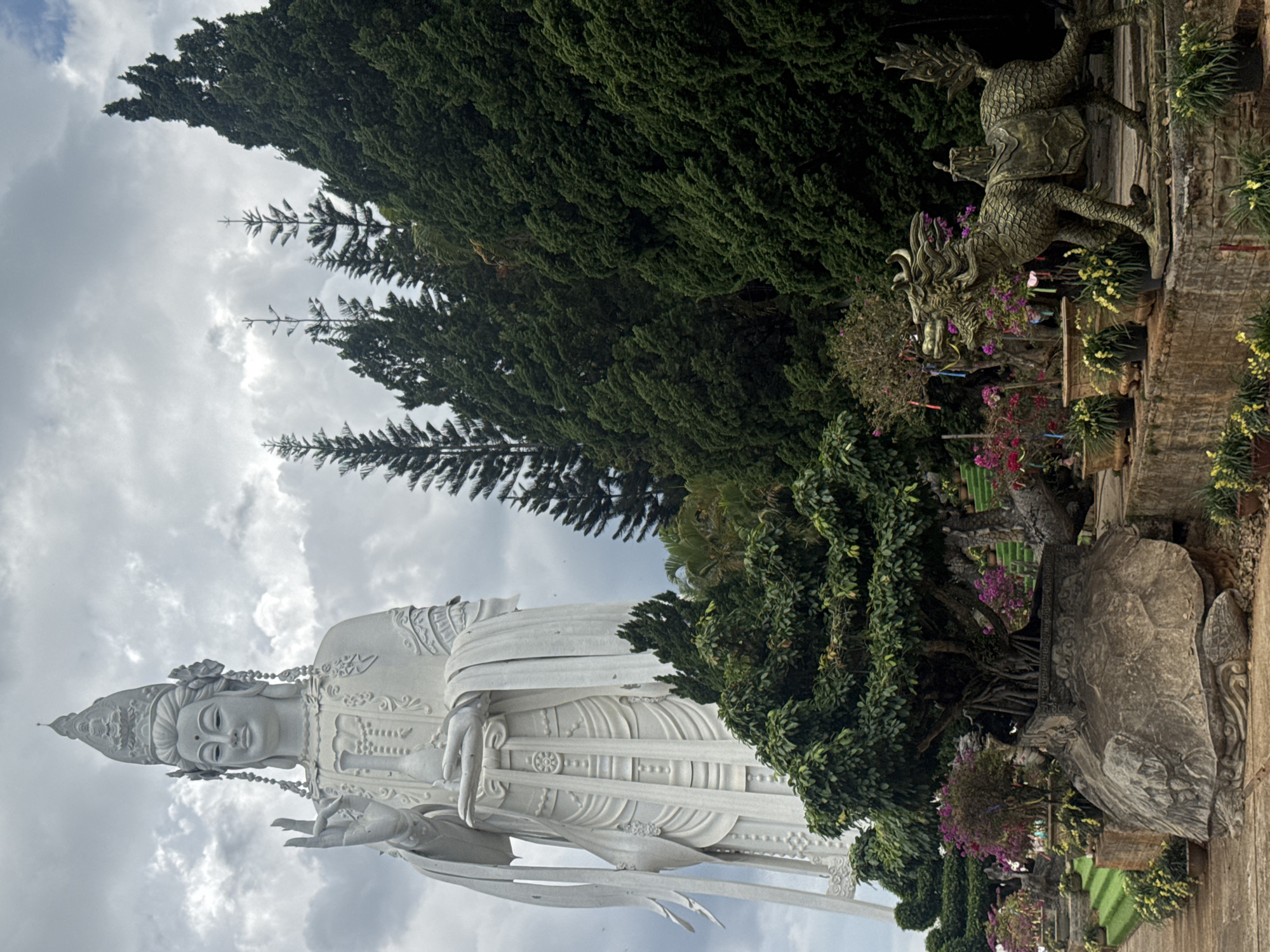 A vertical, eye-level shot shows a large white statue of a deity in the Linh An Pagoda in Nam Ban, Vietnam. The deity, possibly Guan Yin, is standing, wearing robes and a headpiece. To the right of the statue are large green trees and various plants, along with a dragon statue sitting above the plants. The sky is cloudy.