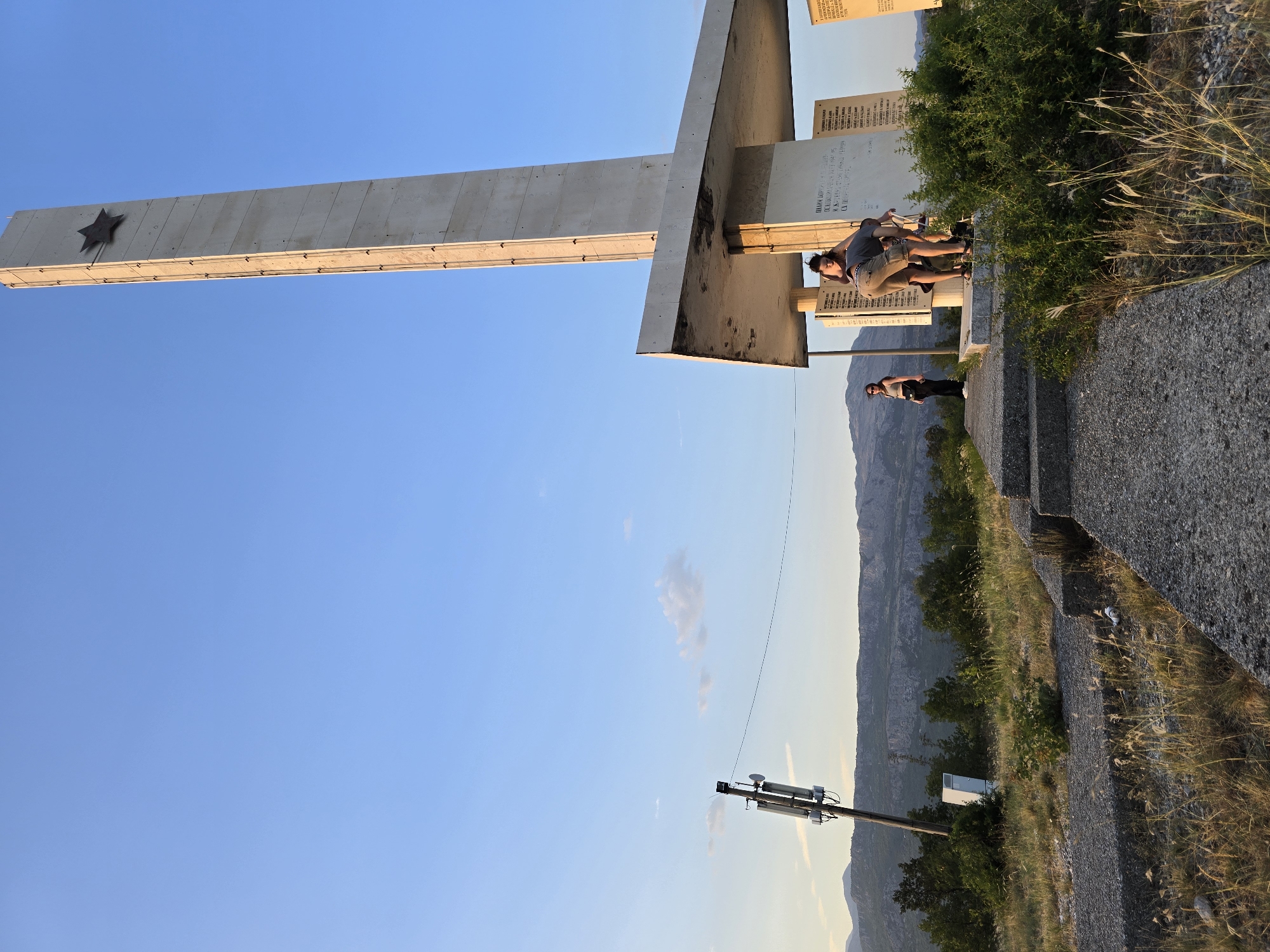 The image depicts the Monument to fallen fighters for freedom, located on the hill of Gorica in Podgorica, Montenegro. The monument is a tall, slender concrete structure topped with a star. At its base, a large concrete platform extends, supported by columns. Two individuals are visible near the base of the monument, one bending down and the other standing. Further to the left, another person stands on a concrete path leading up to the monument. The surrounding environment features dry grass, sparse vegetation, and distant mountains. A cellular tower with antennas is also visible to the left of the monument. The sky is clear blue, with a few faint clouds. The lighting suggests it is late afternoon or early evening.