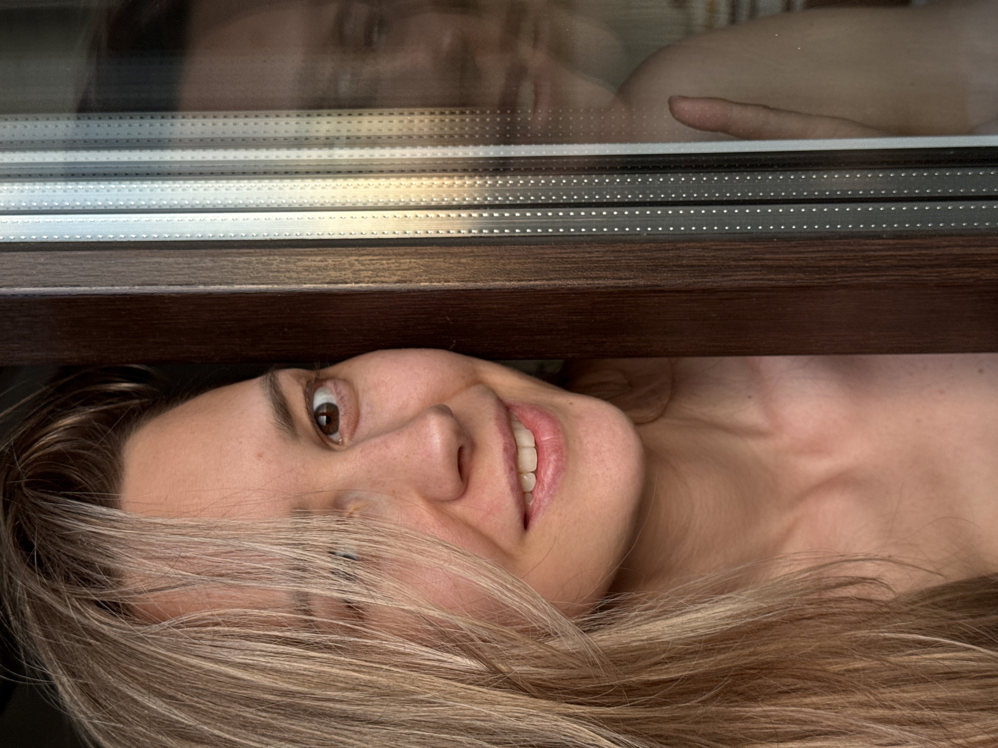 A woman with blonde hair is pictured lying on her side looking up at the camera, her face partially obscured by her hair. Above her is a window frame with a dark wood finish. The window glass shows a blurry reflection of what appears to be a person's head and shoulder. There is no information to verify that the location is in the city of Chaikovsky, Russia.