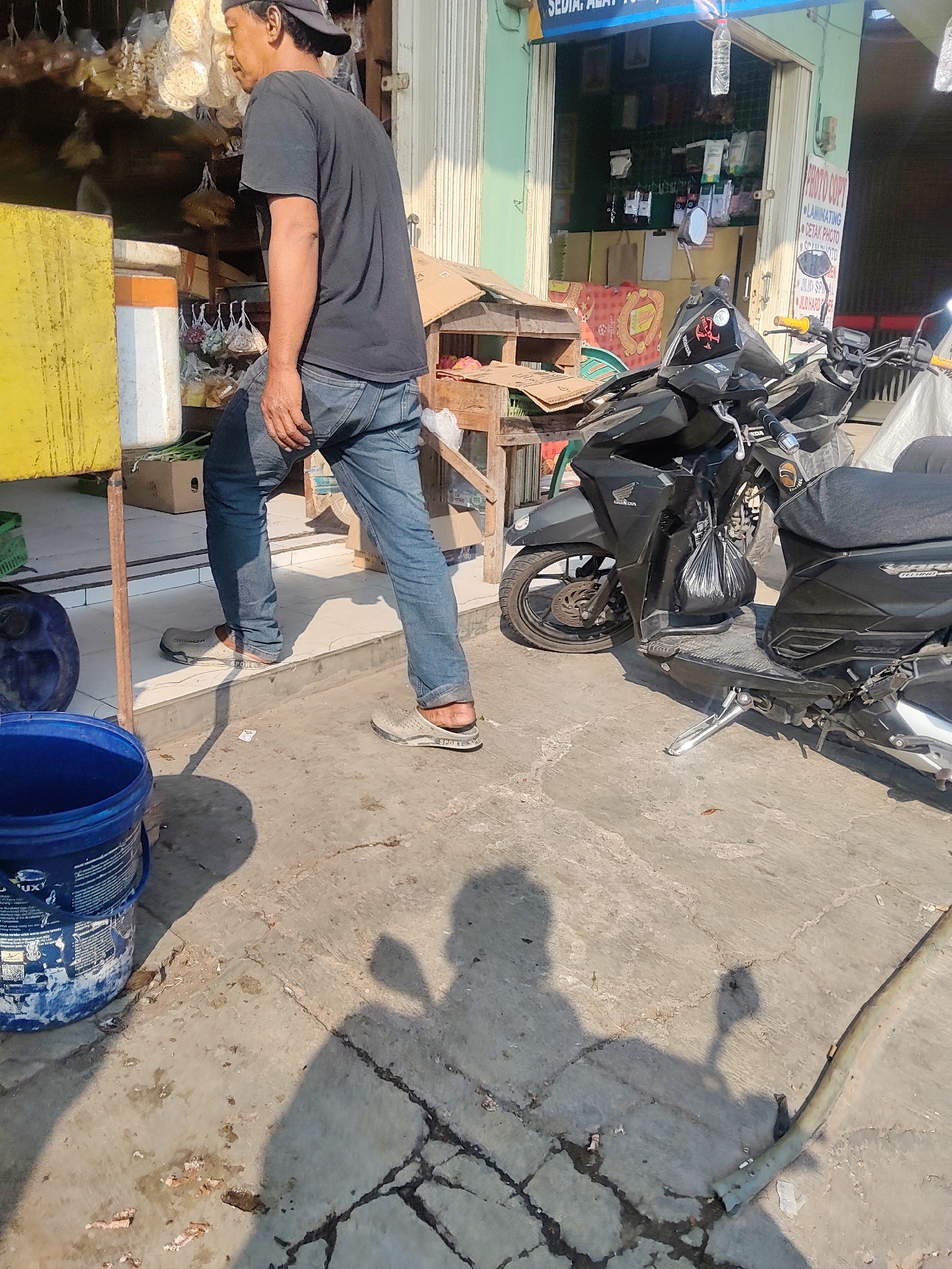A middle-aged man wearing a dark t-shirt, blue jeans, and a baseball cap is walking past a storefront in Mekarmukti, Indonesia. He is wearing open-backed sandals. To his left is a yellow rectangular object, a white and orange cylindrical object, and a blue plastic bucket. Behind him, the storefront displays various items for sale, including bags of dried goods hanging from the ceiling. A black motorcycle is parked in front of the storefront. The ground is paved and cracked, with scattered debris. The scene is lit by bright sunlight, casting shadows on the ground.
